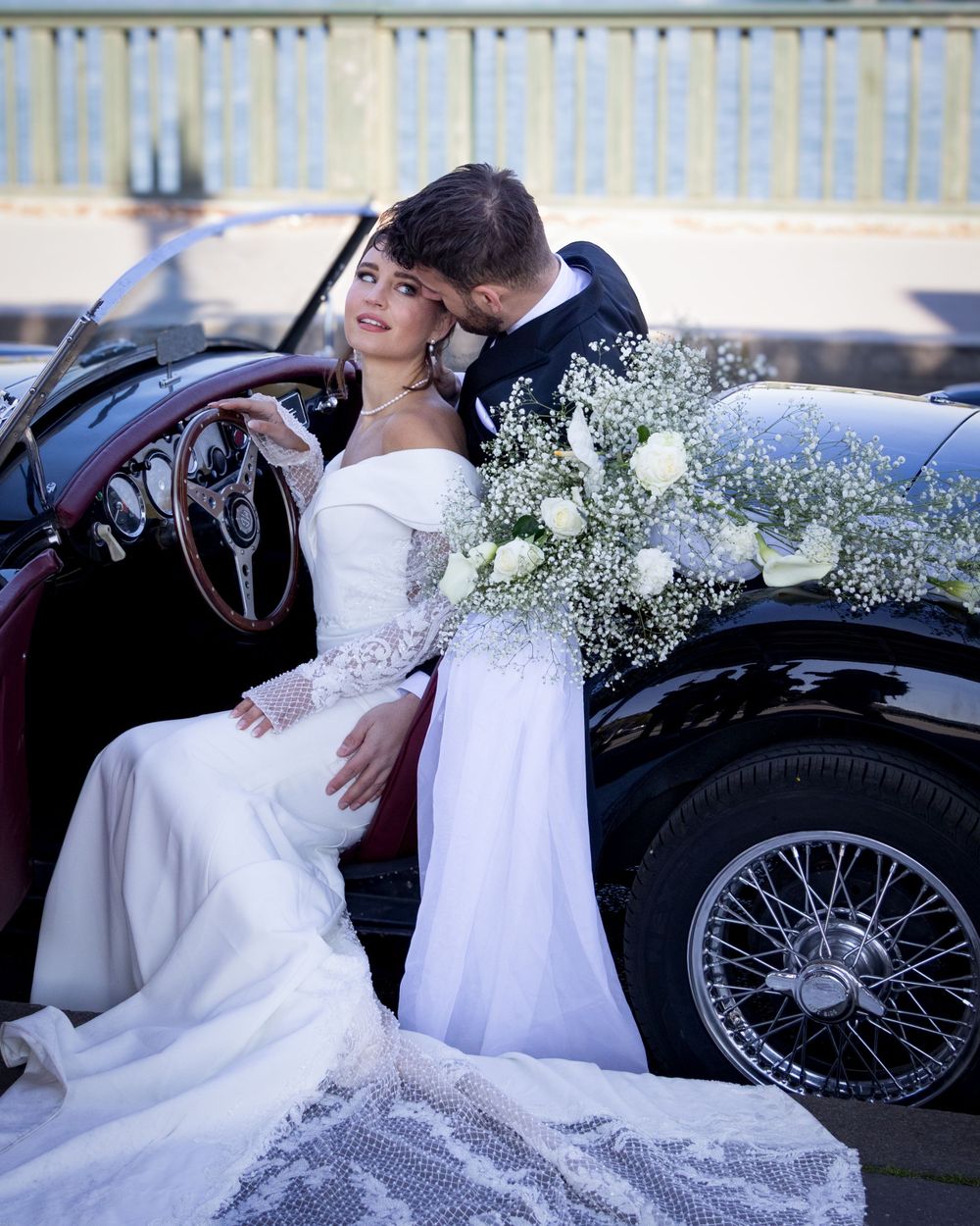 Couple in wedding attire pose romantically in vintage classic car with white flowers and delicate lace details.