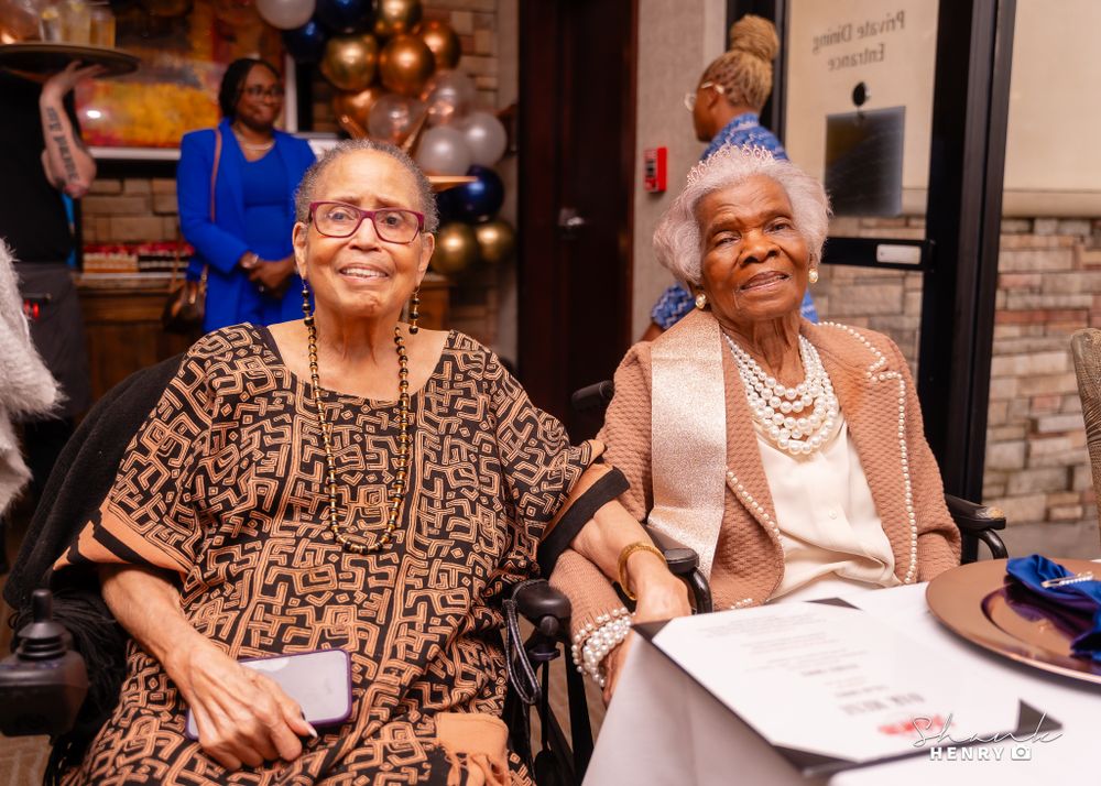 Two seniors wearing elegant outfits smile while seated at a celebratory event.