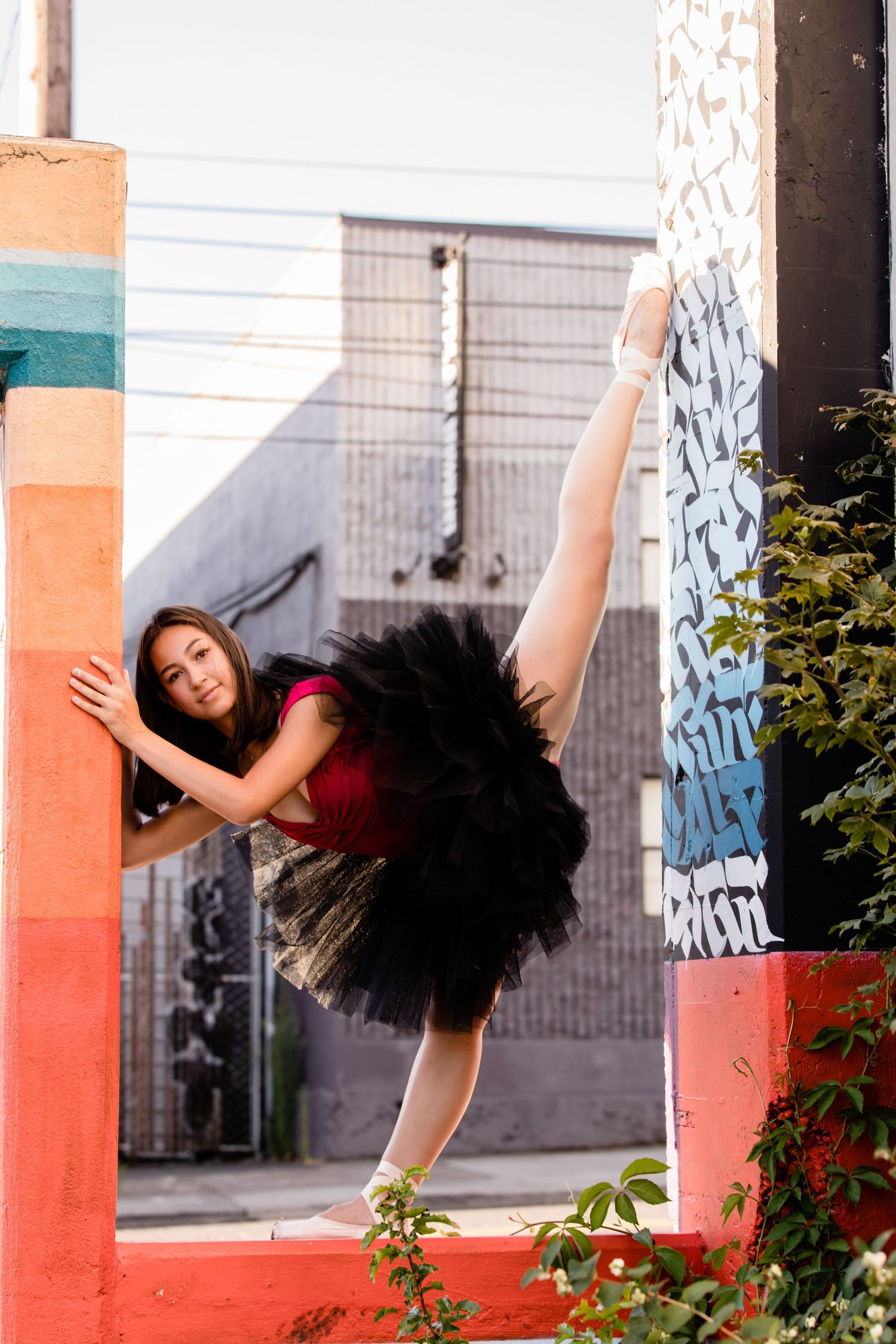 A dancer in a red top and black tutu performs a graceful extension against a colorful urban wall with graffiti.