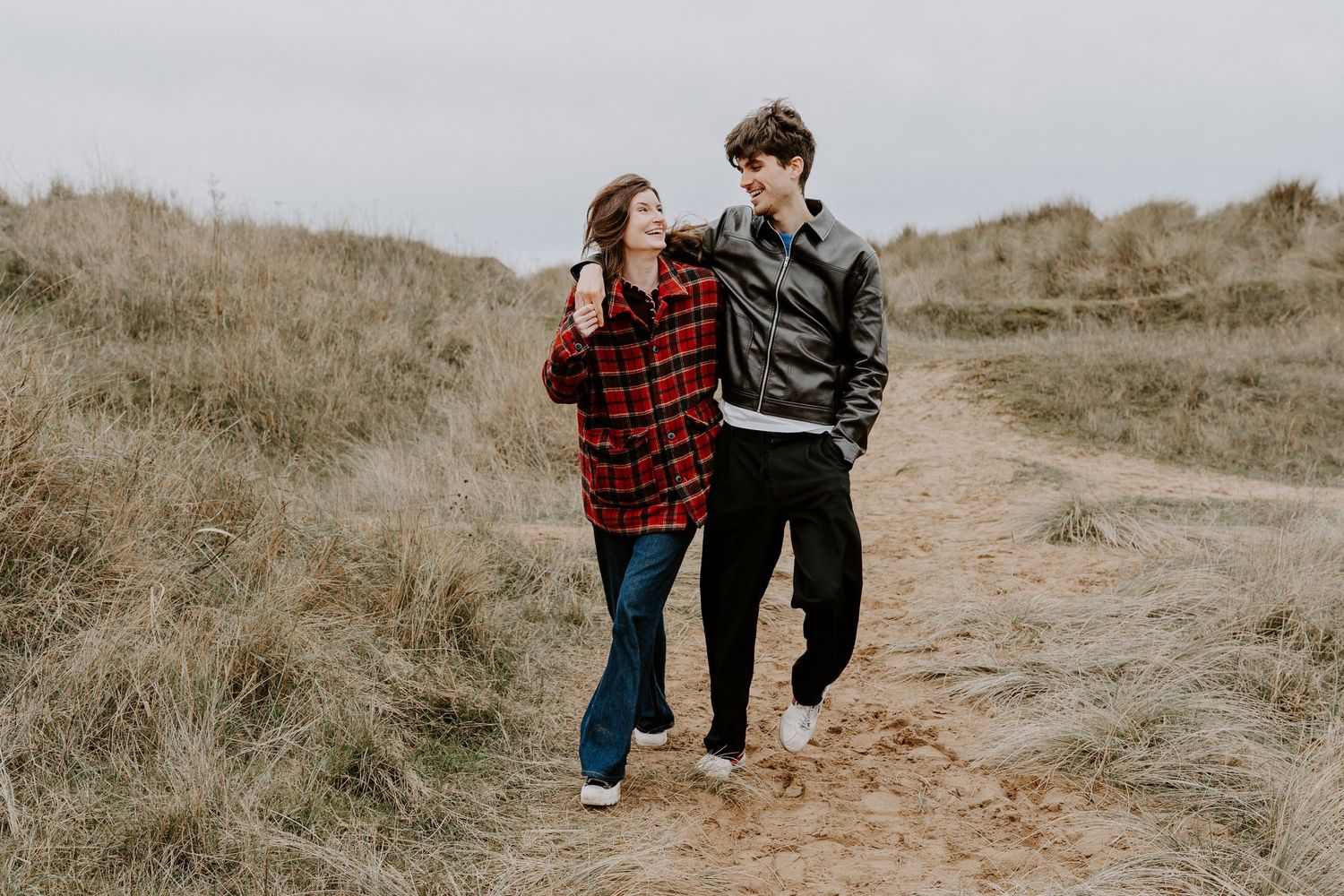 A couple walking on the beach. The man has his arm draped round the ladies shoulder. they are looking at each other and smiling.