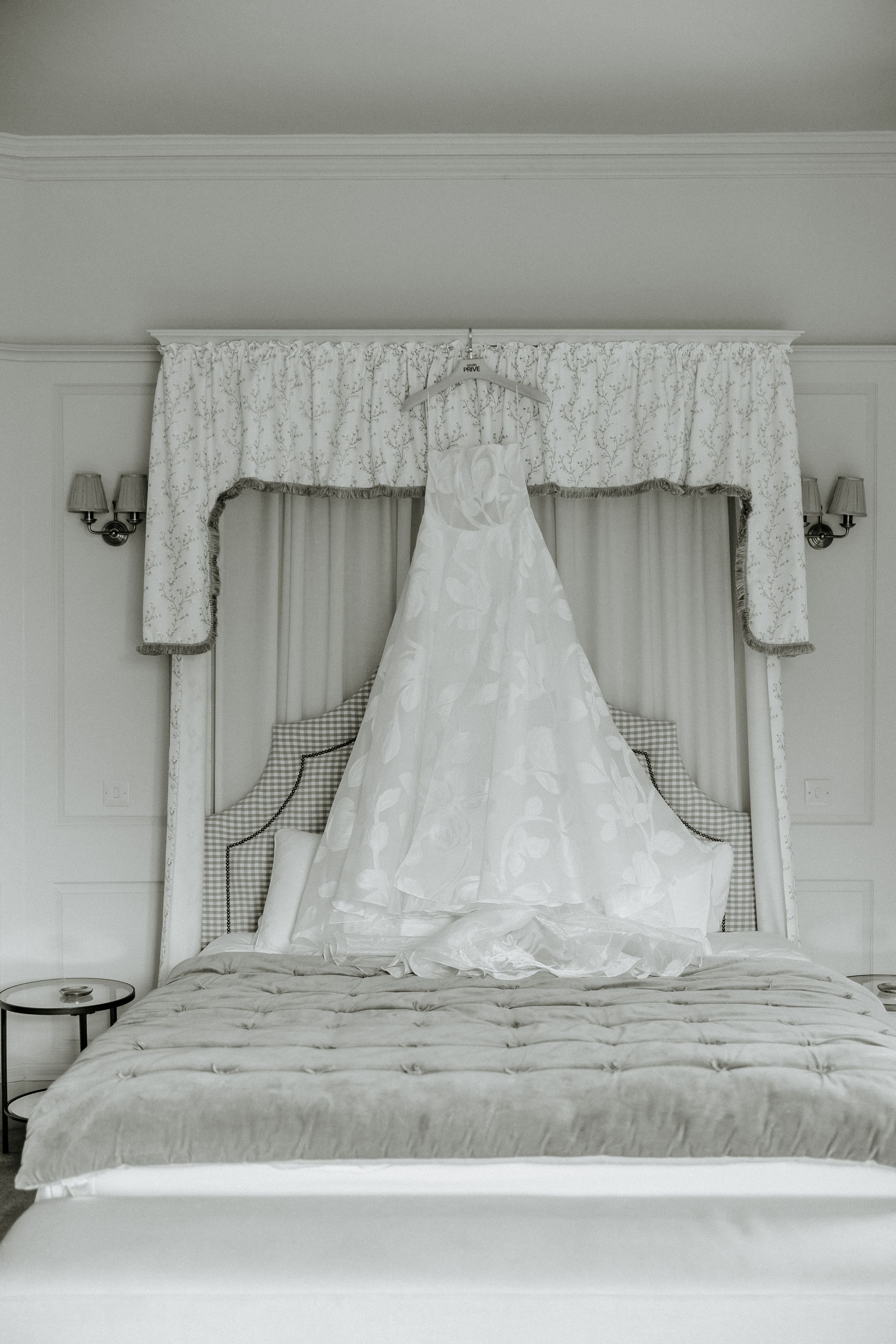 Elegant white lace wedding dress hanging displayed against gray curtains in a formal bedroom setting.