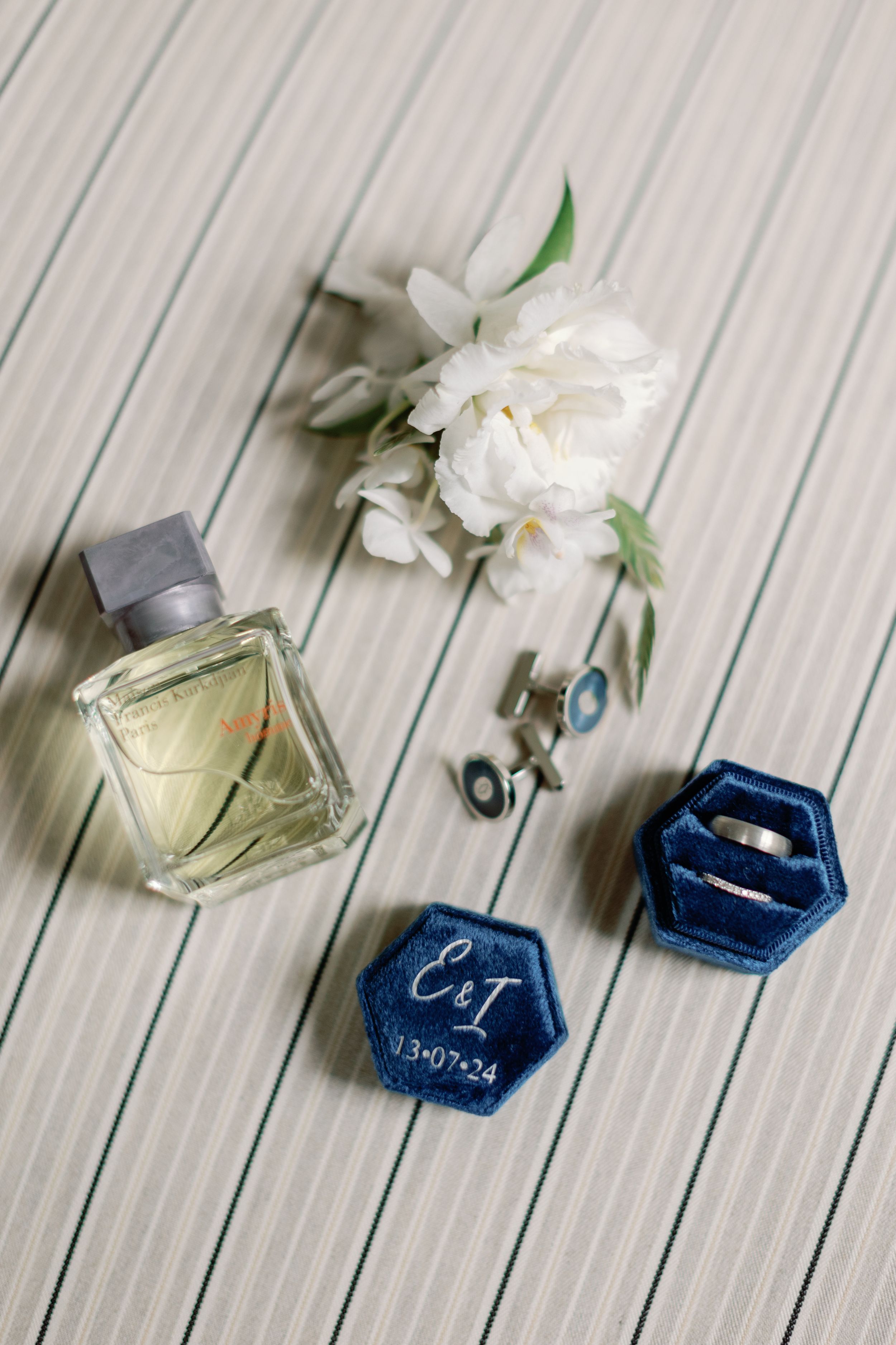 Wedding details including perfume bottle, white flowers, rings and blue velvet ring boxes on a striped surface.