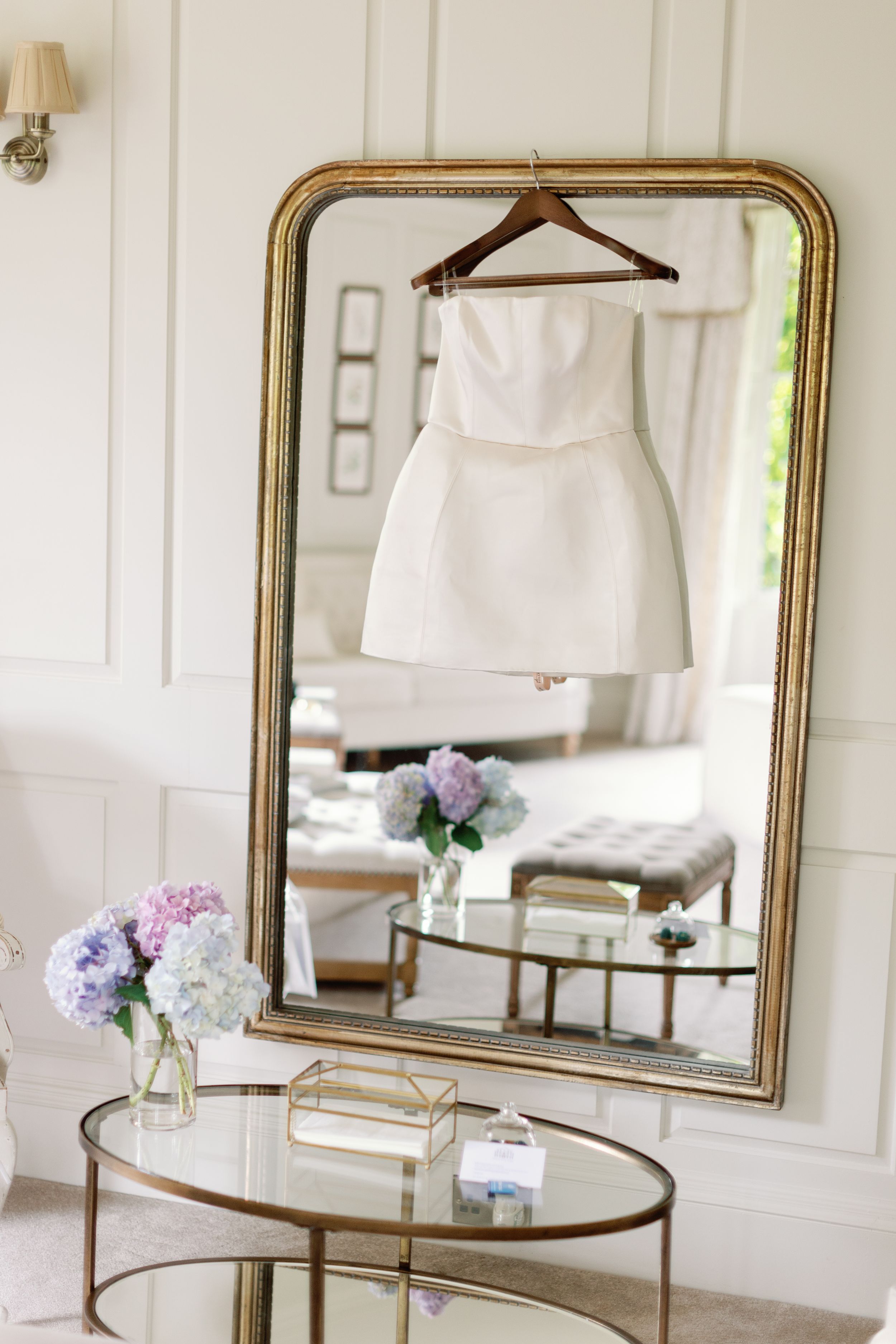 White formal dress reflected in gold-framed mirror above glass coffee table with pastel flowers.