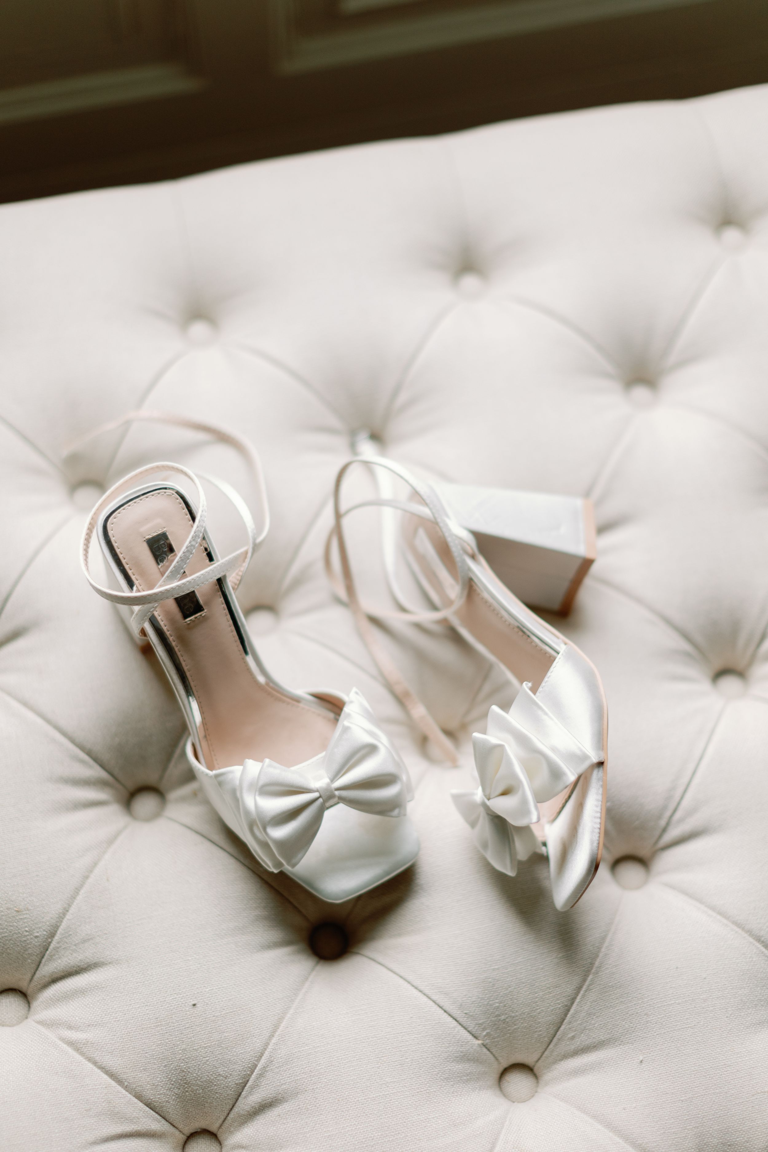 Elegant white satin wedding shoes with bows displayed on a tufted light gray cushion.
