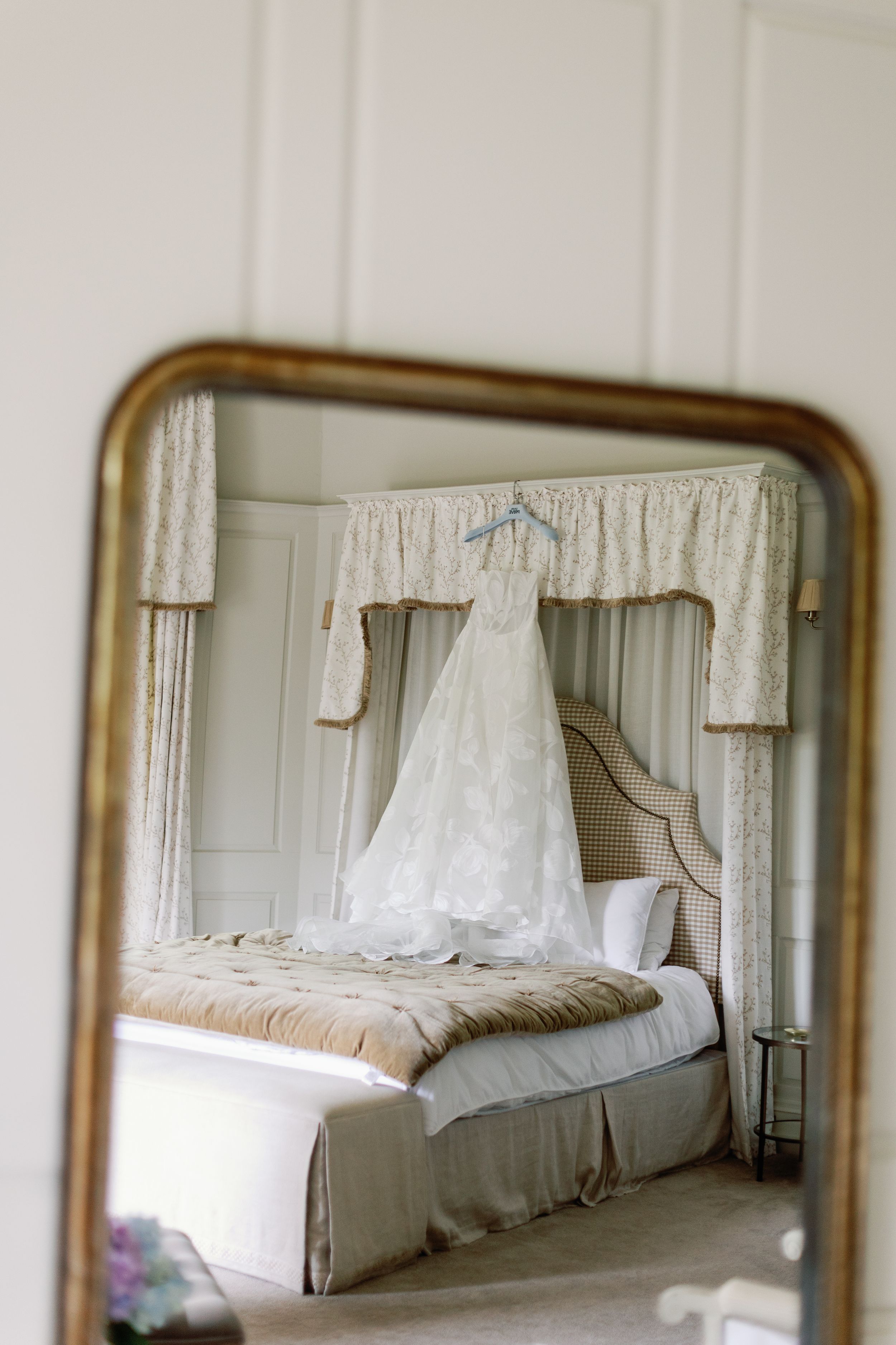 A white lace wedding gown hangs above a bed in a bedroom, reflected in an antique gold-framed mirror.