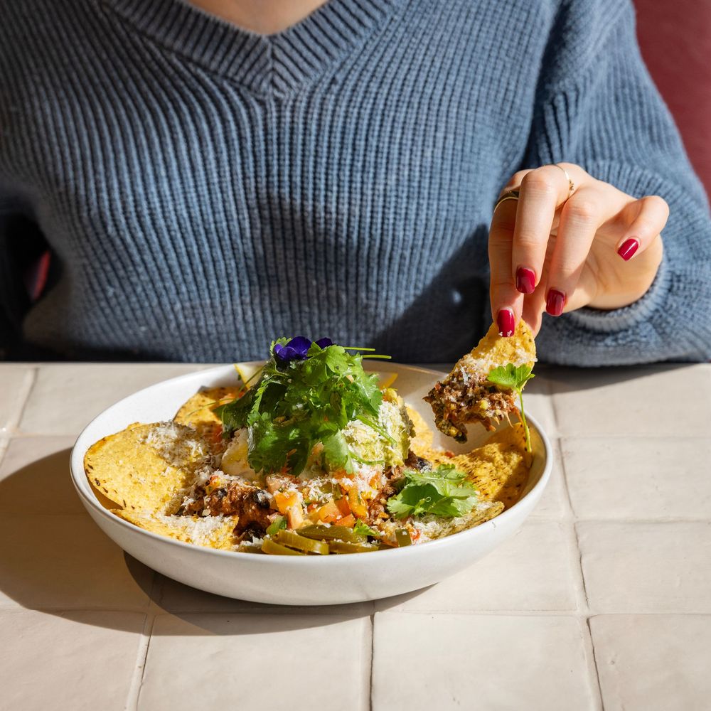 Nachos sitting on a white tile background with a woman in a blue top and red fingernails holding some