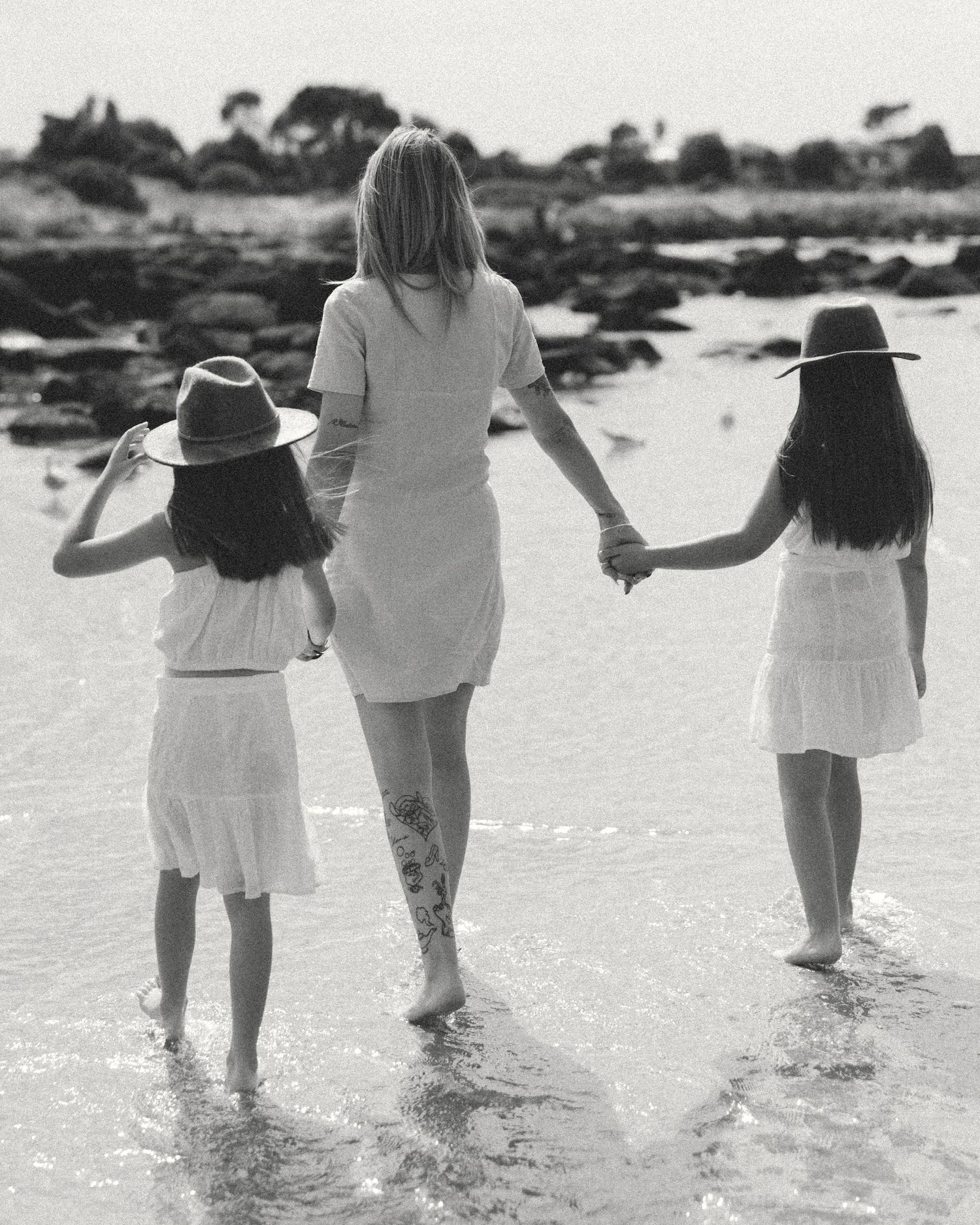 Black and white beach scene showing three people holding hands while walking along the shore in white dresses.