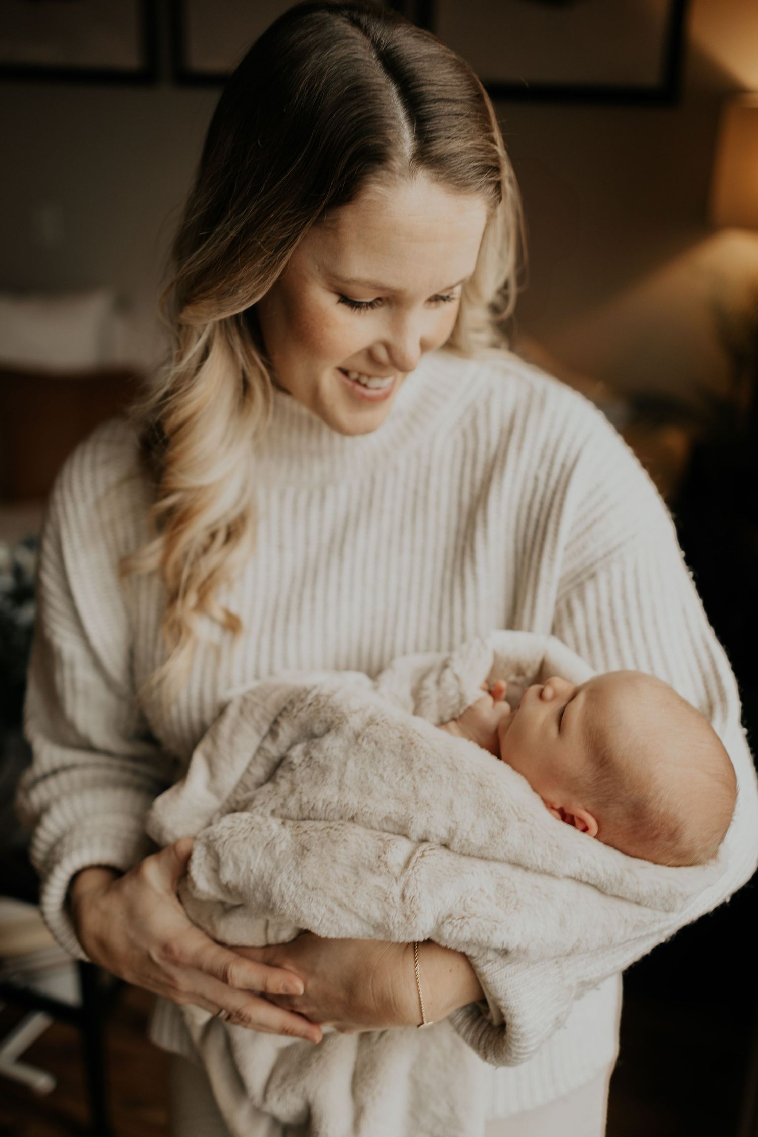 A person in a cozy cream sweater gently holds a sleeping newborn baby wrapped in a soft blanket.