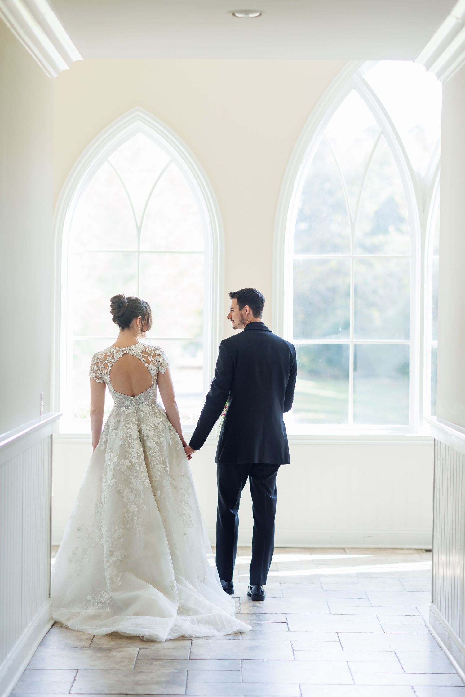 A couple hold hands walking down a bright church hallway with arched gothic windows and white walls.