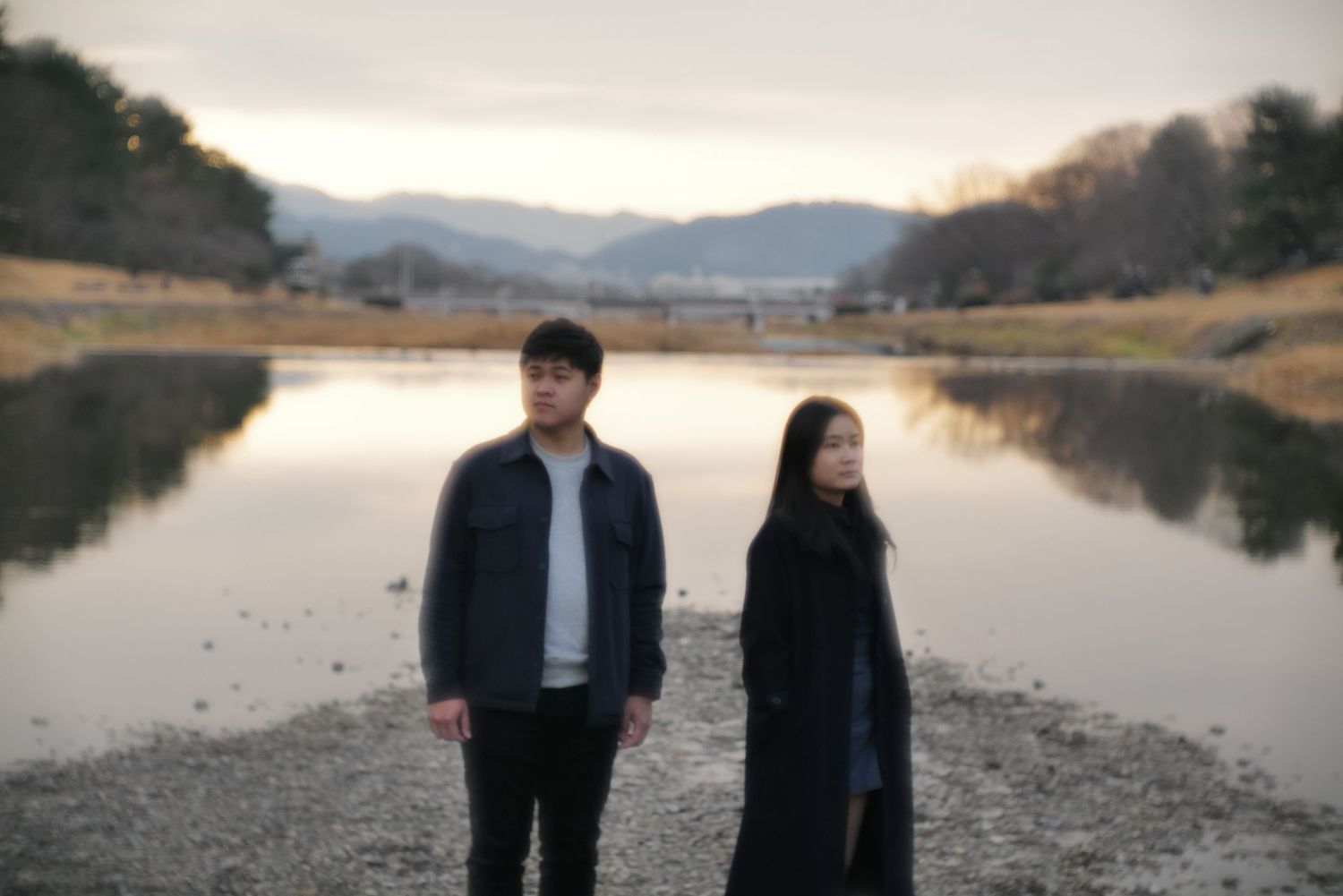 Young couple standing apart near a calm river with mountain backdrop at dusk.
