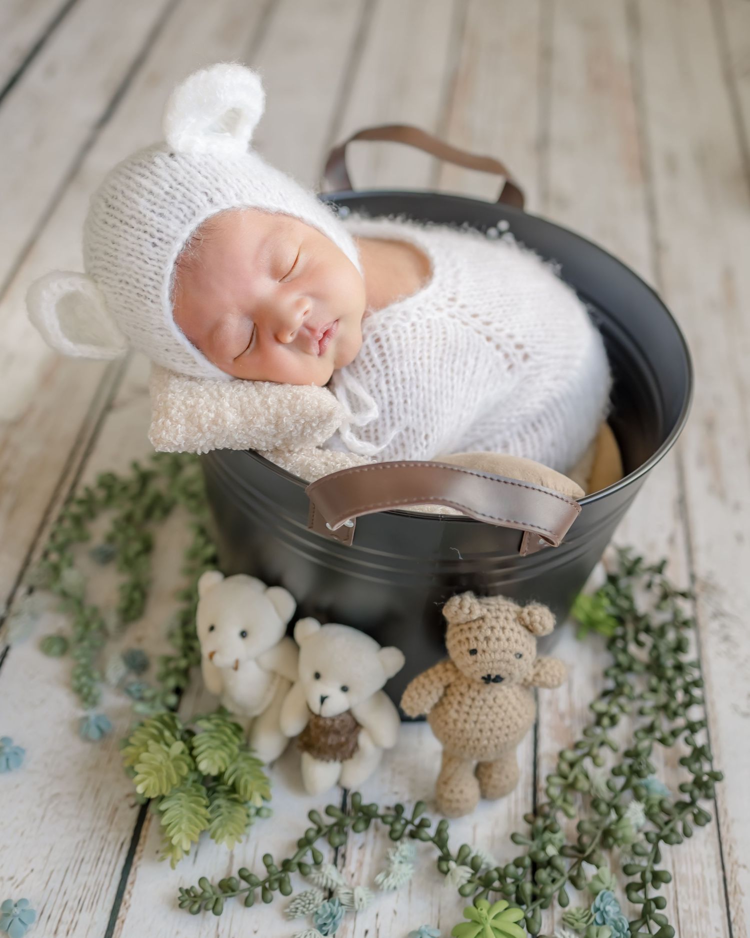 Sleeping newborn in white knit outfit nestled in metal bucket with small teddy bears and greenery on wooden floor.