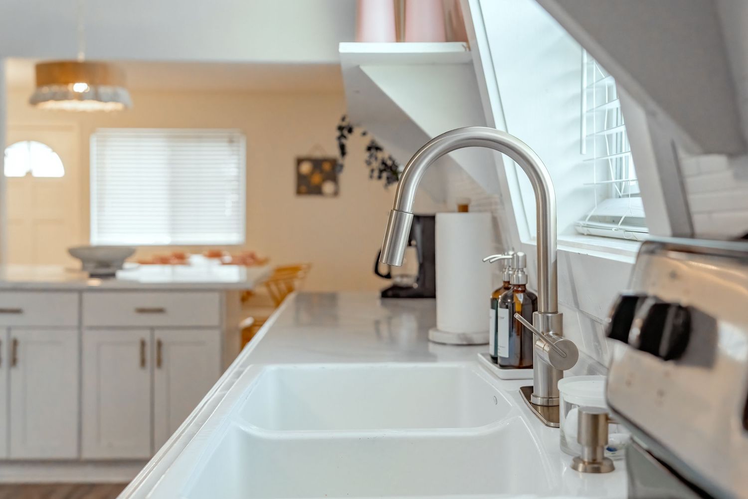 Modern white kitchen interior with farmhouse sink and stainless steel faucet against white quartz countertop.