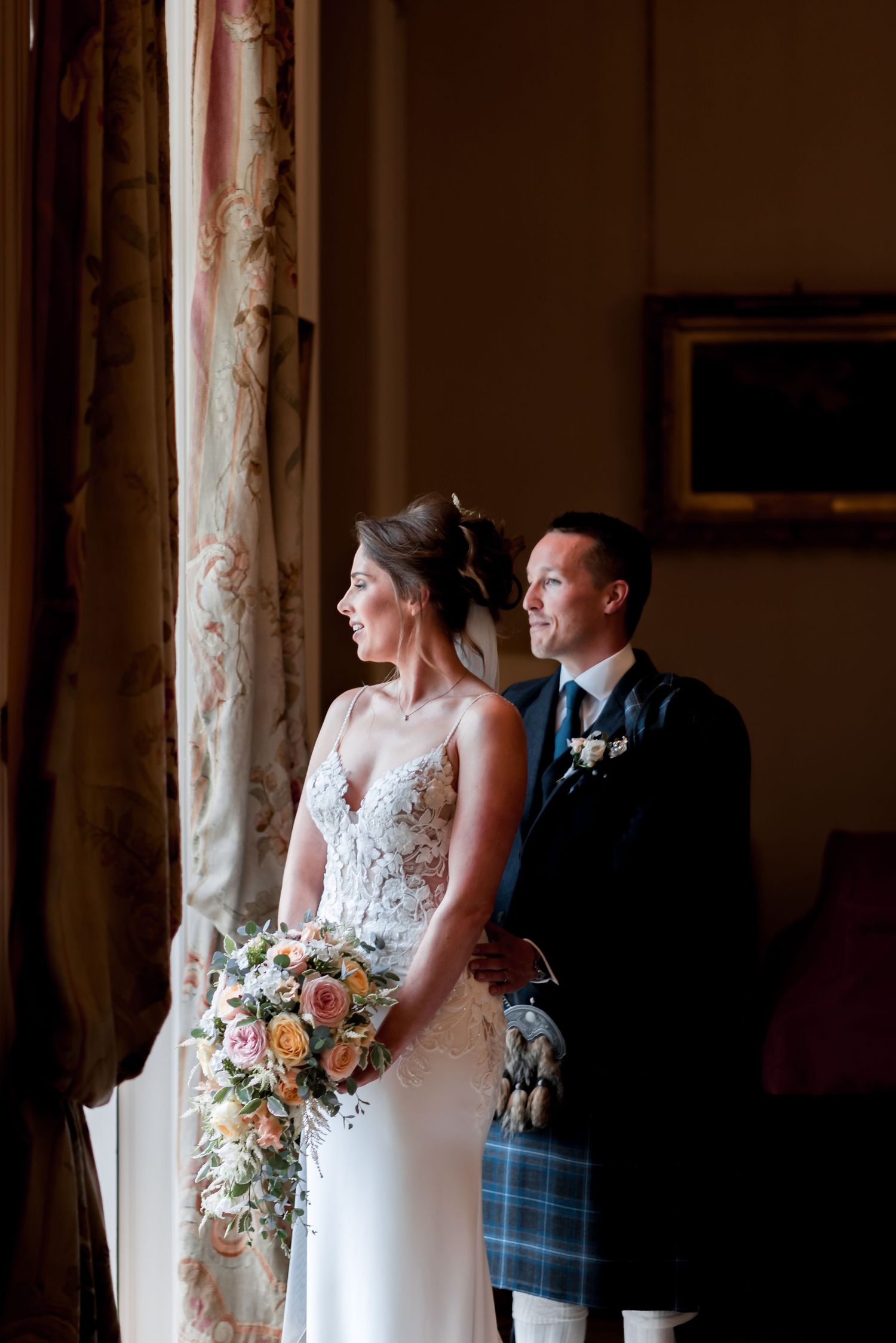 Couple in wedding attire poses by window, with bride in lace dress holding peach floral bouquet and groom in Scottish kilt.