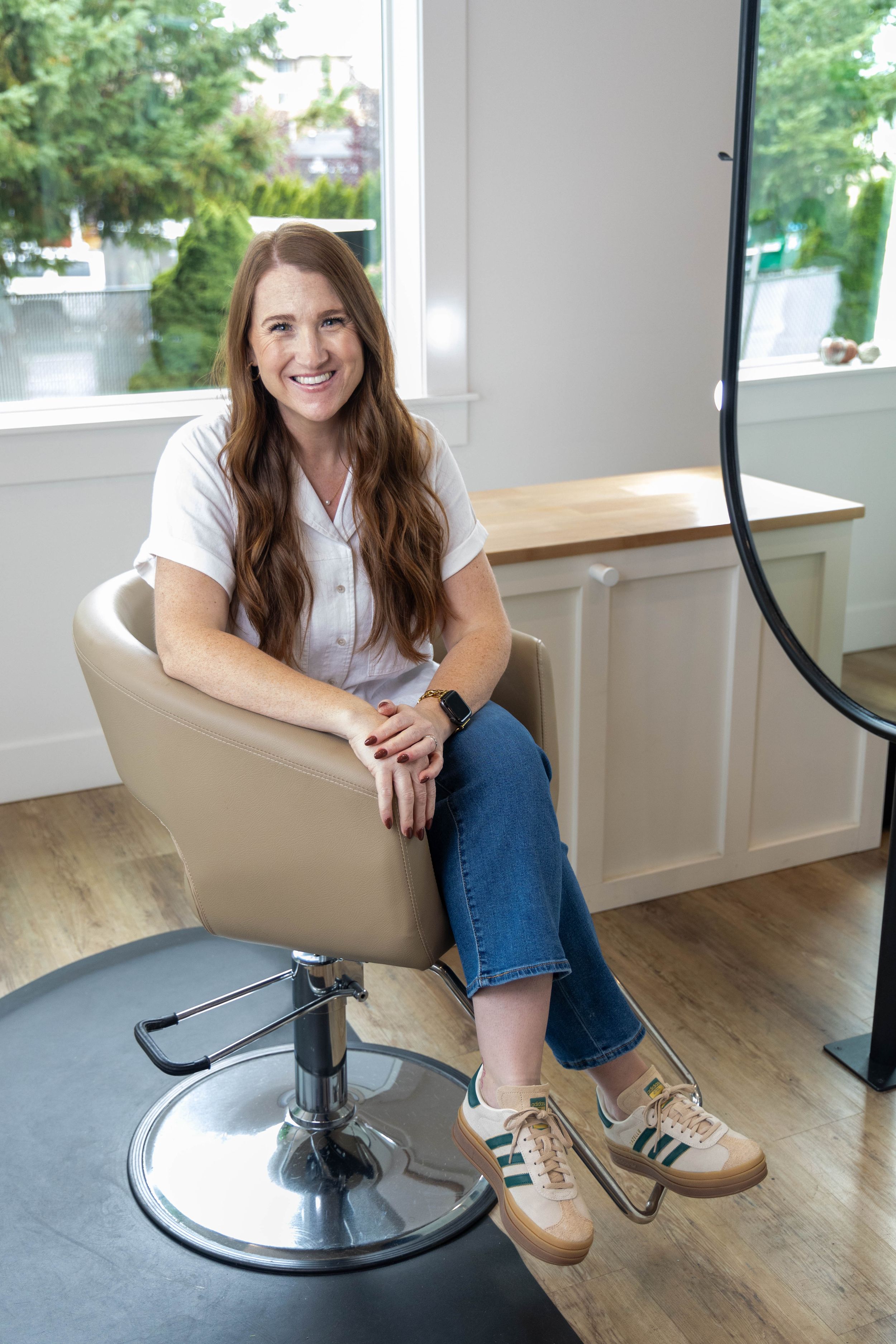 A salon professional seated in a tan styling chair wearing jeans and a white top in a modern studio space.
