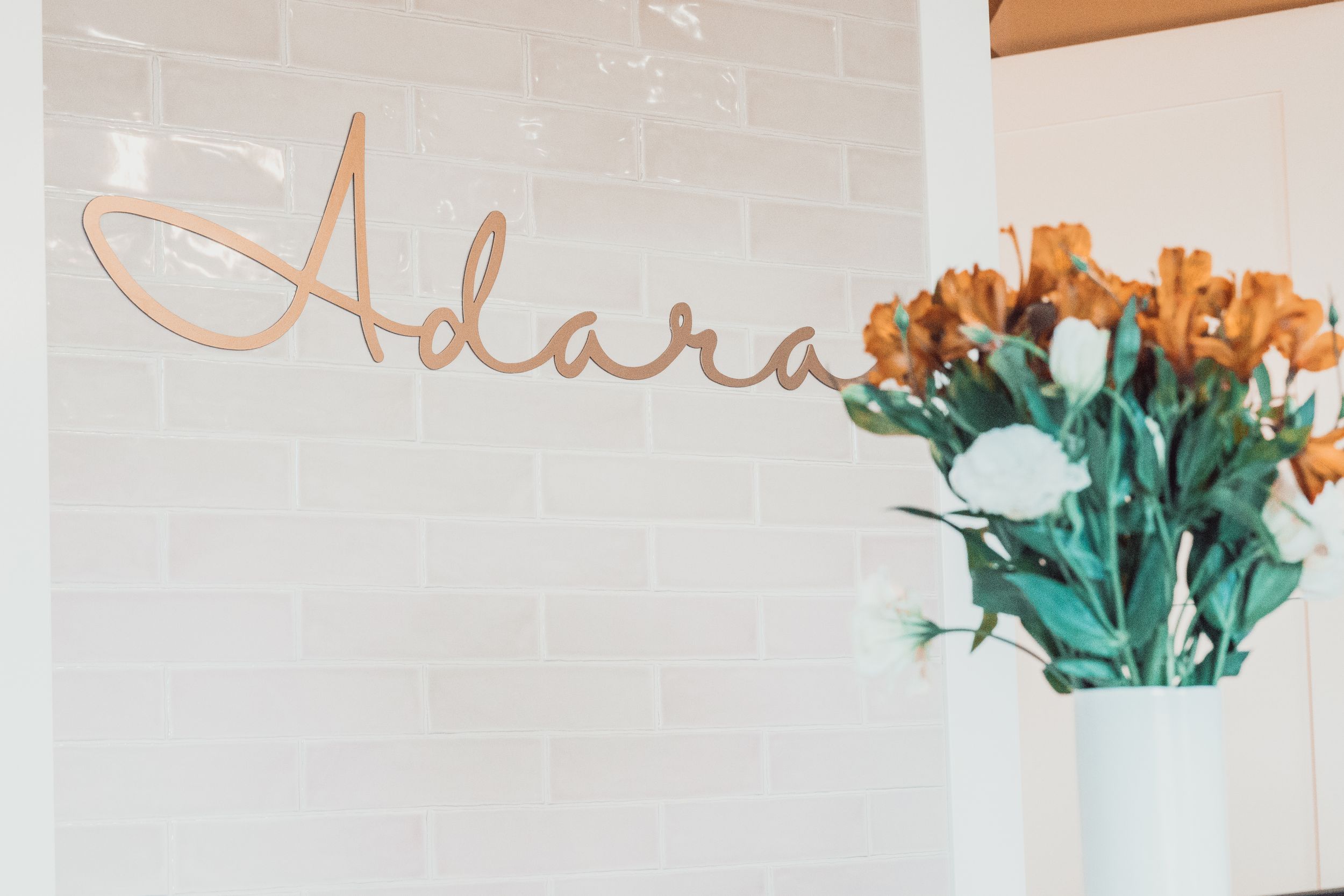 An elegant cursive name 'Adara' written on a wall beside a vase of orange roses with green stems.
