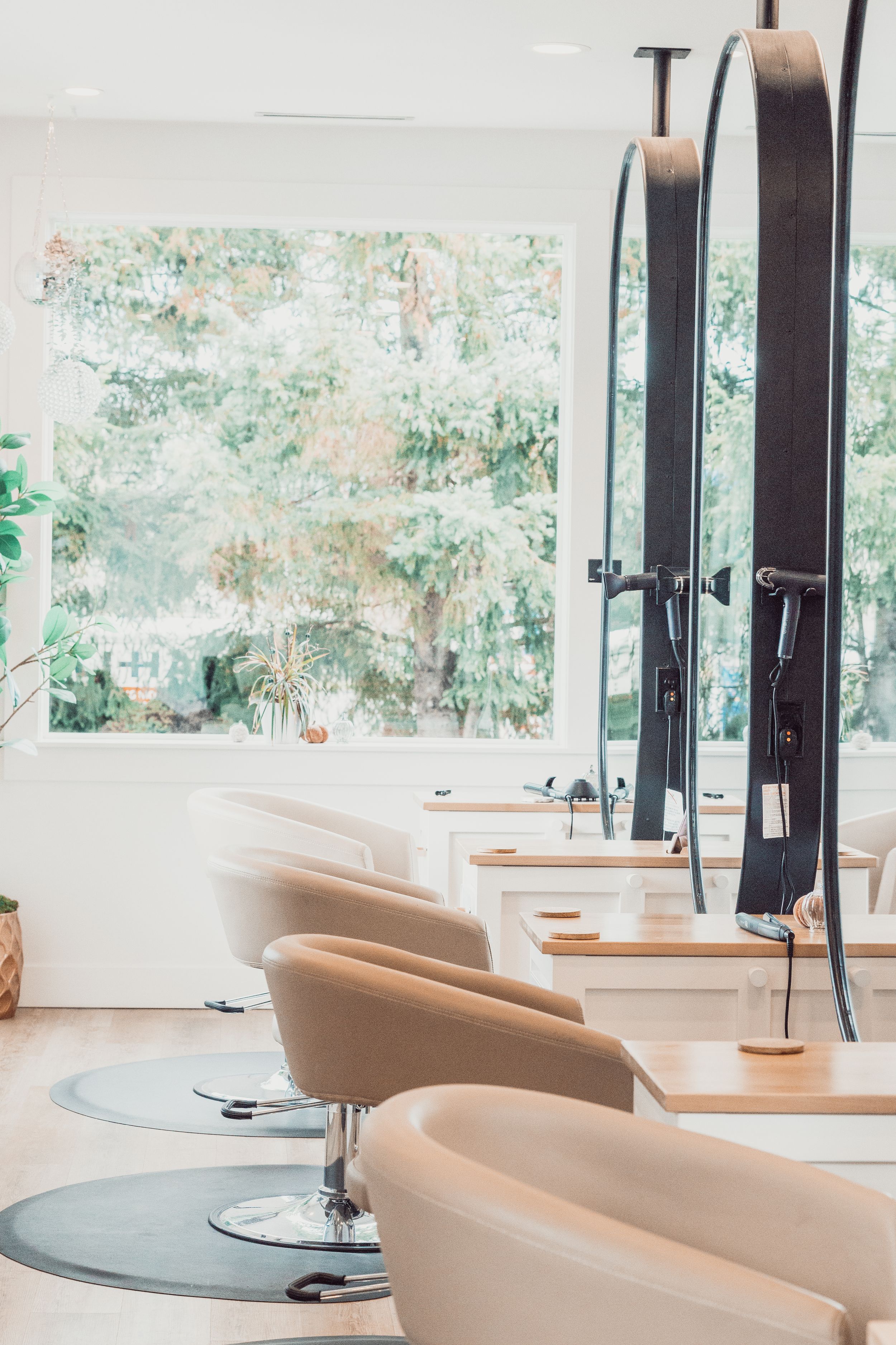 Modern hair salon interior with beige swivel chairs and large window overlooking lush greenery.