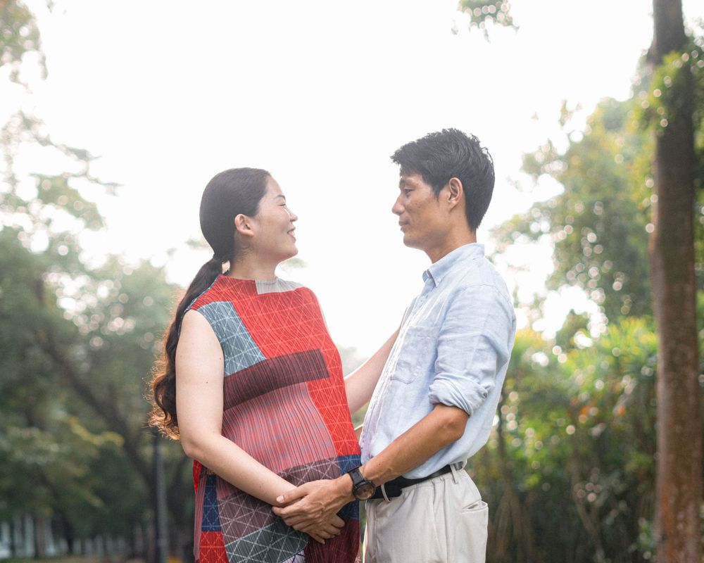 An expectant couple in casual attire shares a tender moment outdoors in a natural setting.