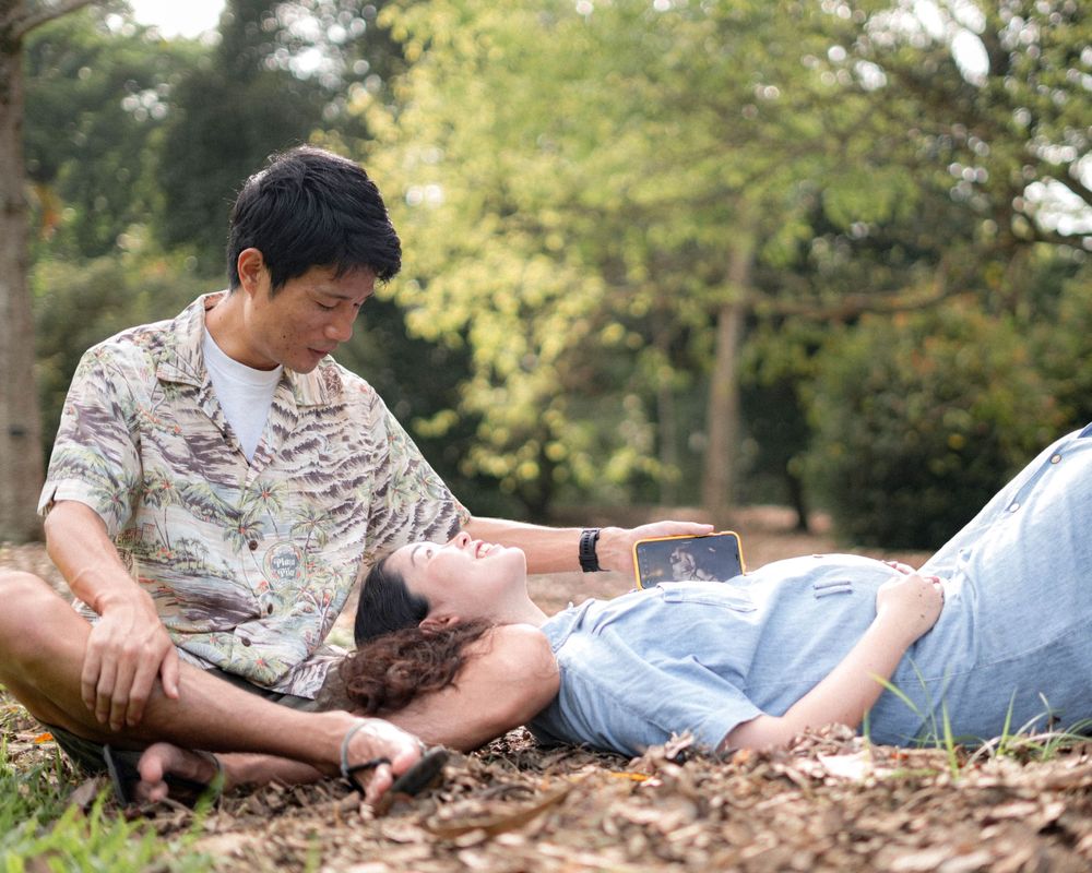 A relaxed maternity photoshoot scene with a couple resting on fallen leaves.