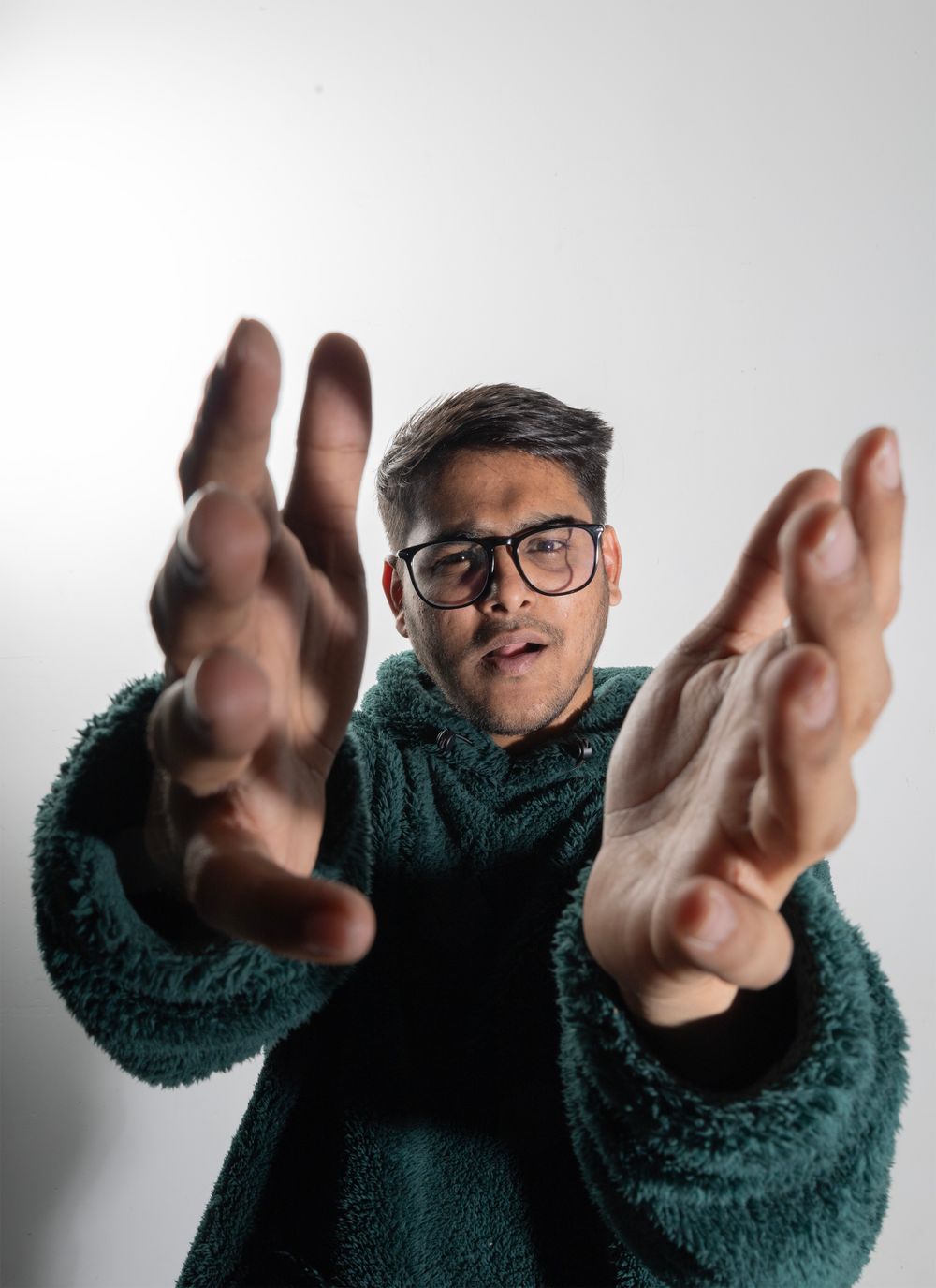Person in teal fuzzy sweater and glasses reaching toward camera with dramatic hand gestures.