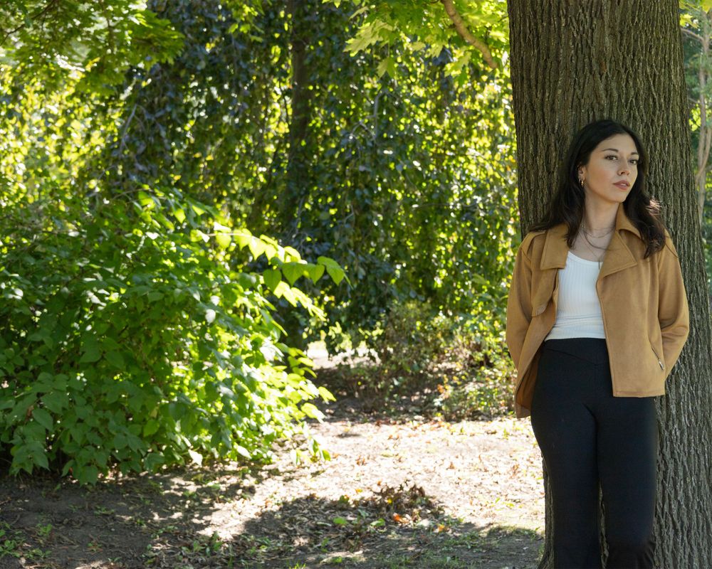 Casual outdoor portrait of someone in tan jacket and black pants leaning against tree.