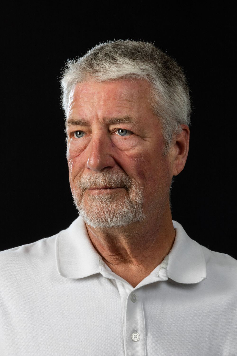 Studio portrait of a senior man in a white polo shirt against dark background.