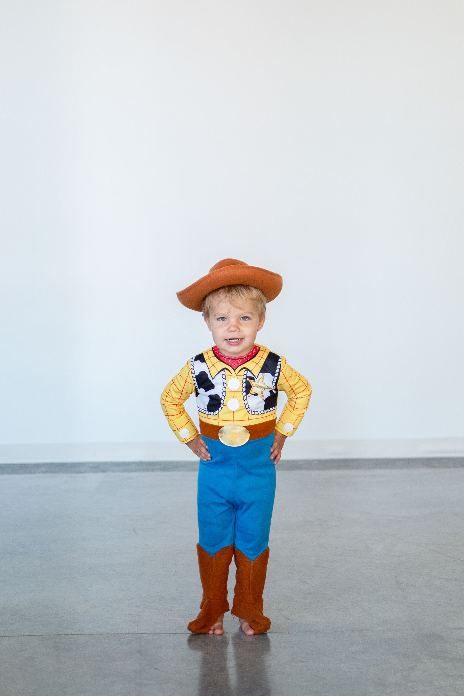 A child dressed in a Woody costume from Toy Story stands against a gray wall with hands on hips.