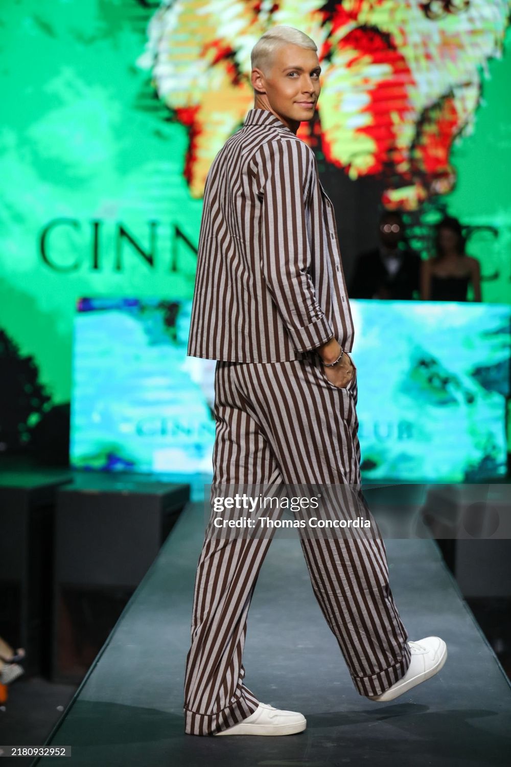 A model walks the runway wearing a gray and white striped matching set with white sneakers against a green backdrop.