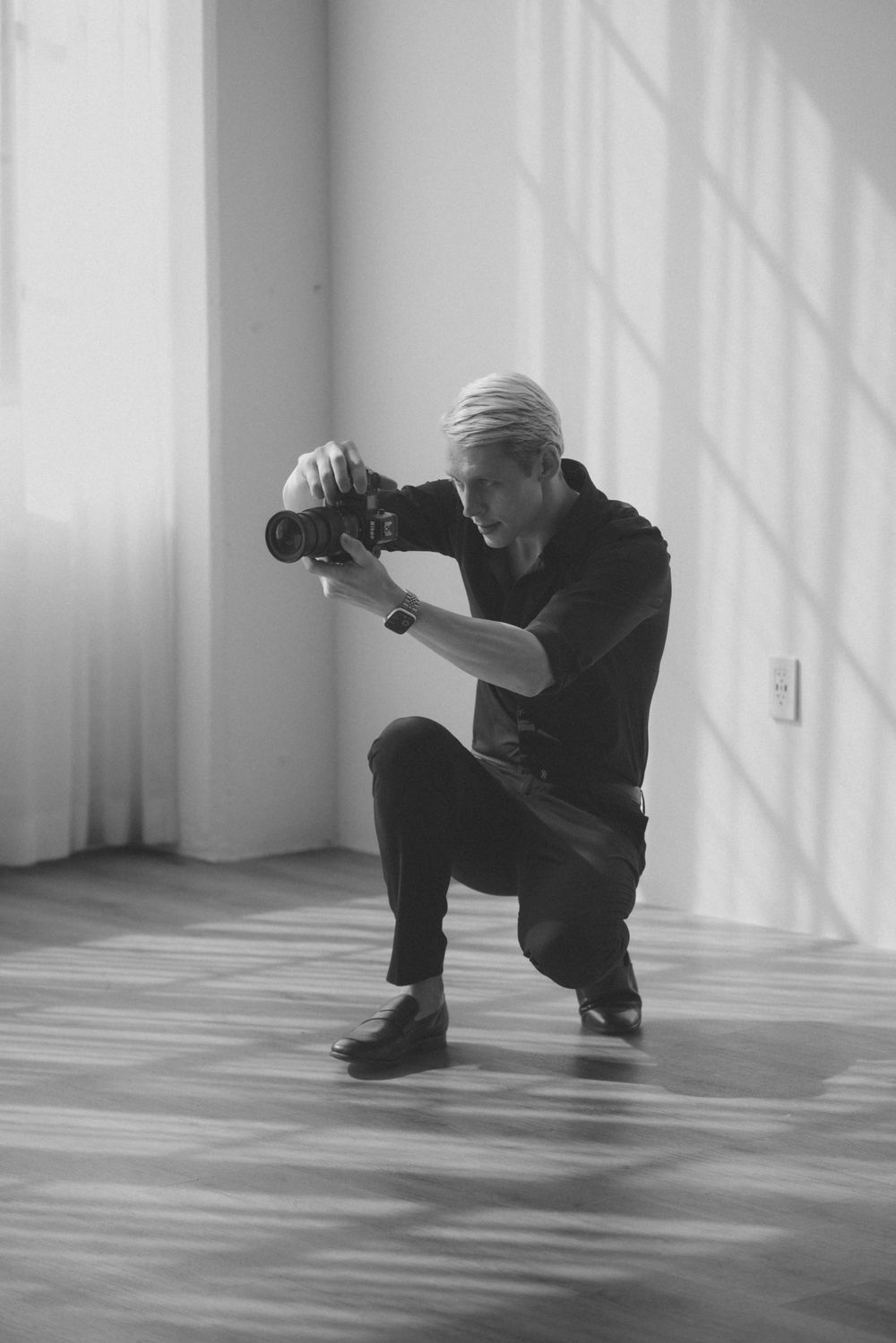 A photographer in dark clothing crouches low to capture shots from multiple angles in a minimalist white room.