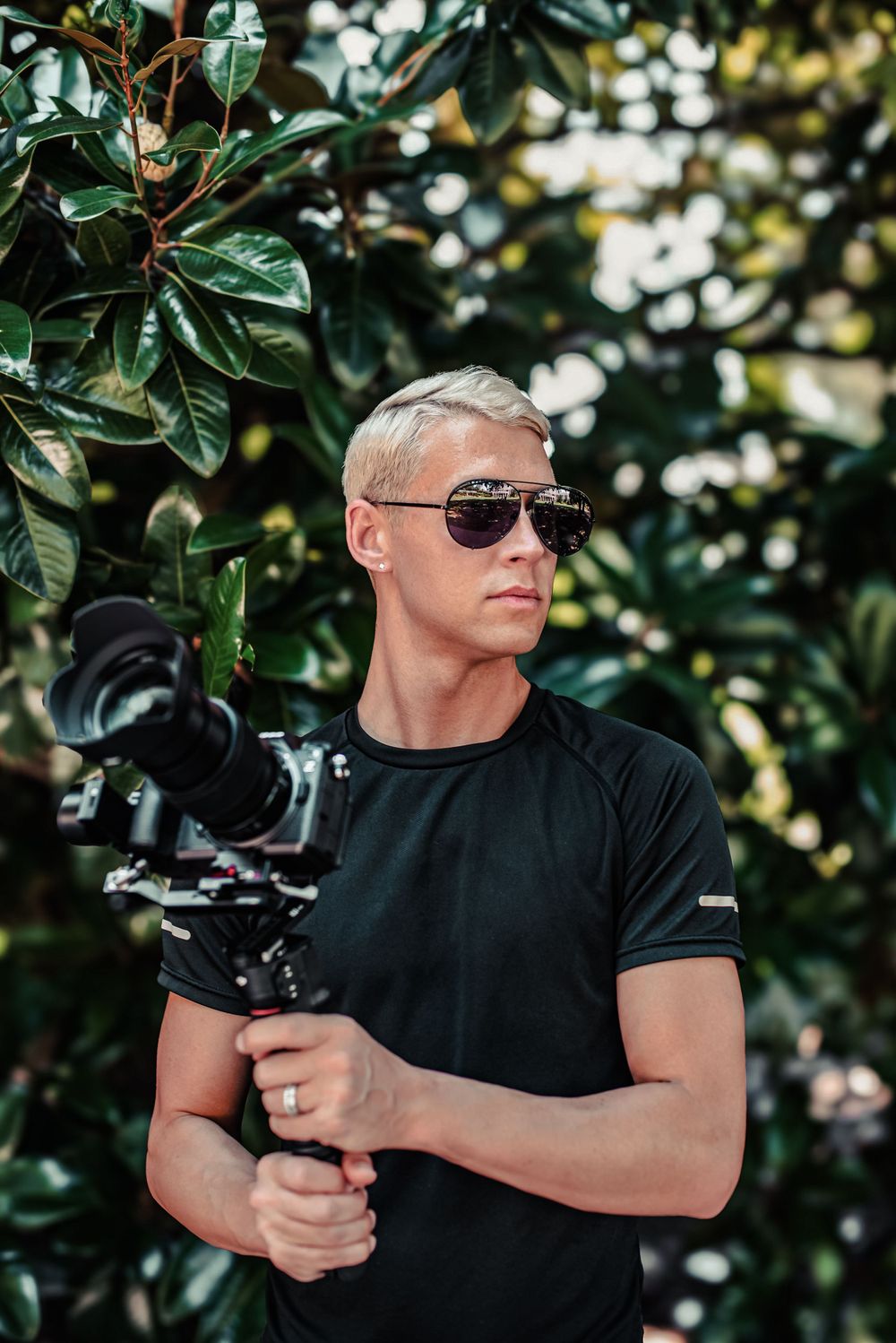Professional videographer in black t-shirt holding stabilized camera equipment against green foliage background.