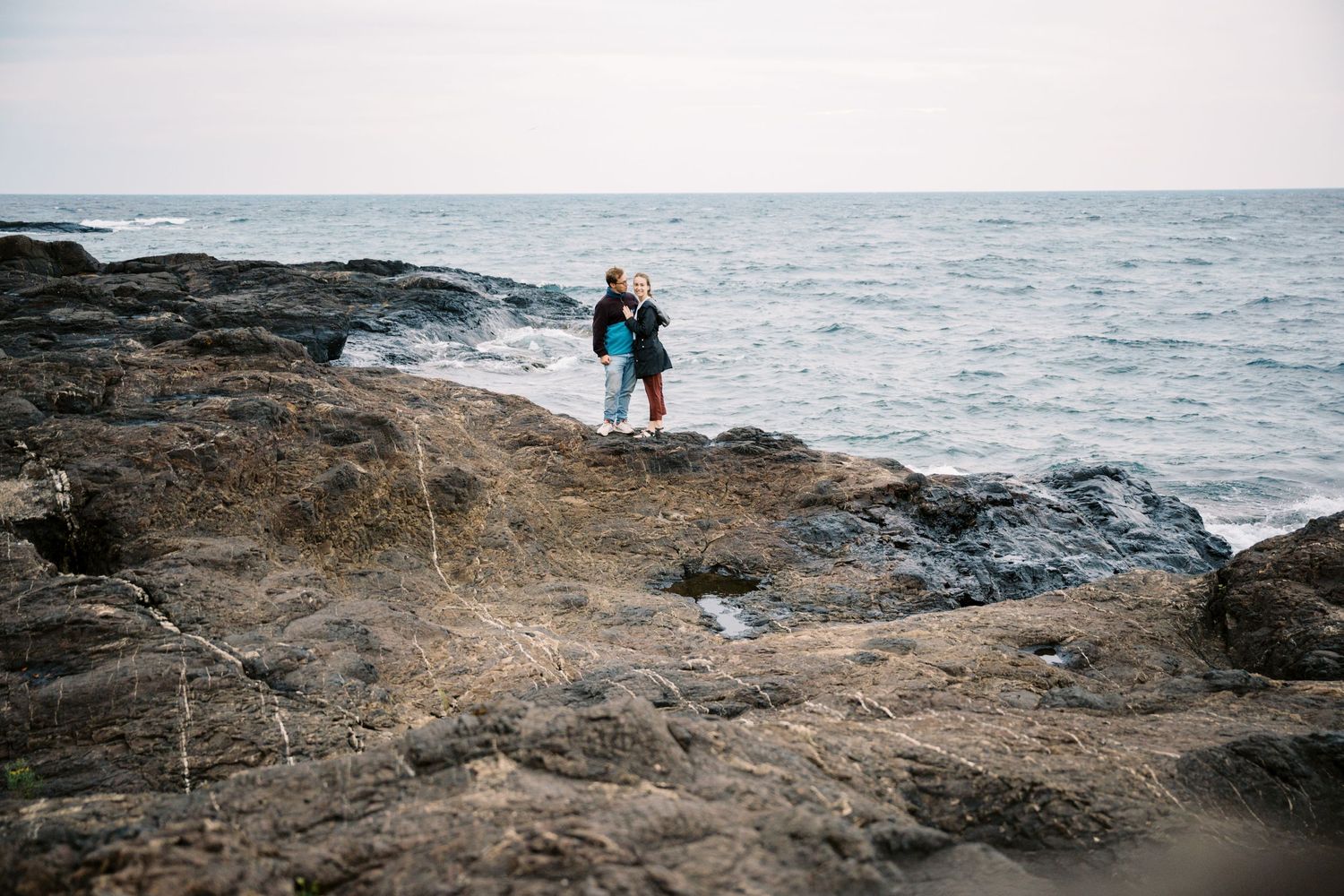 Person standing on rocky coastal outcrop overlooking stormy ocean waves during overcast weather.