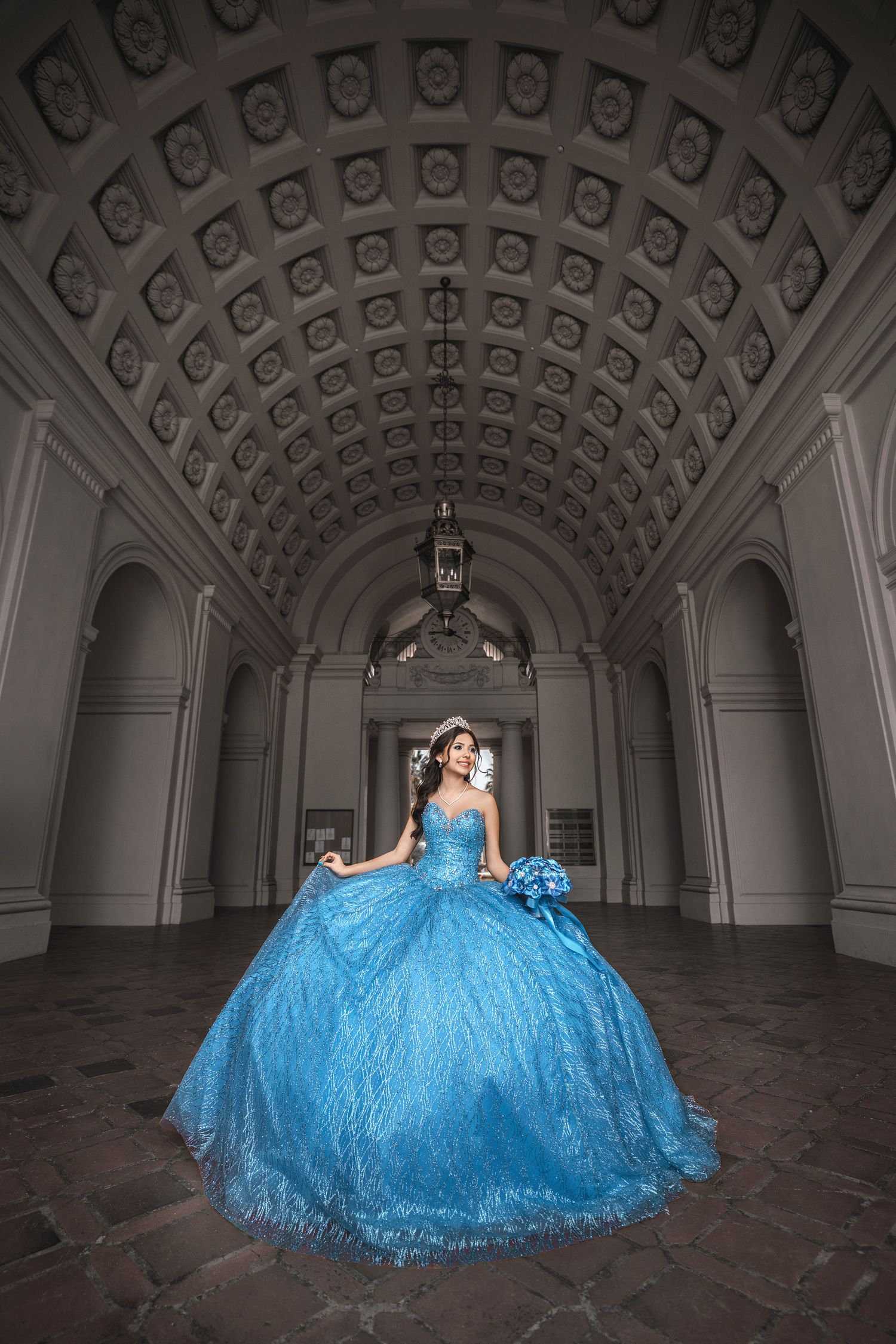 A person in a voluminous sparkly blue ball gown poses beneath an ornate arched ceiling with decorative coffers.