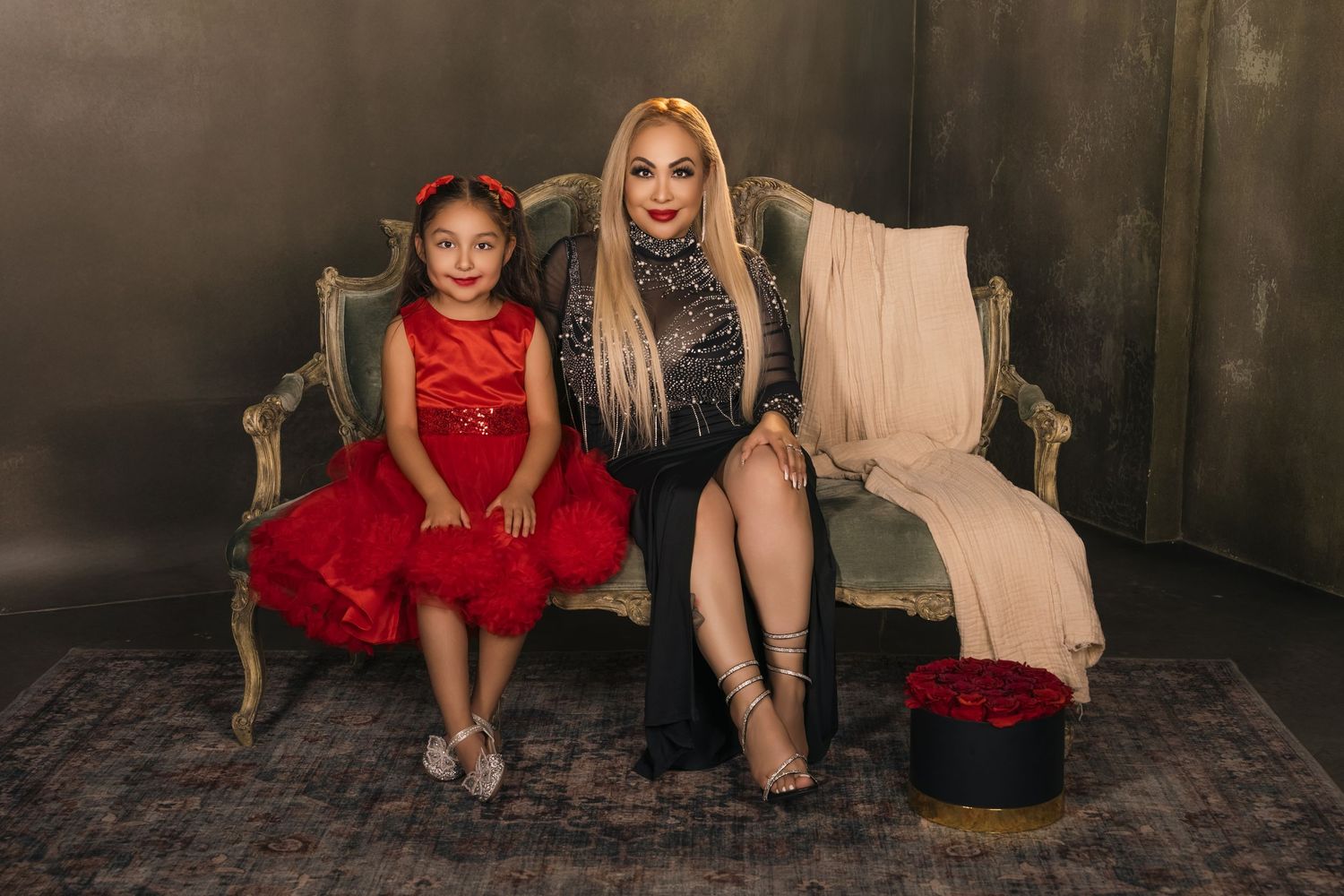 Mom and daughter sit together on an elegant vintage couch, with one wearing a vibrant red dress.