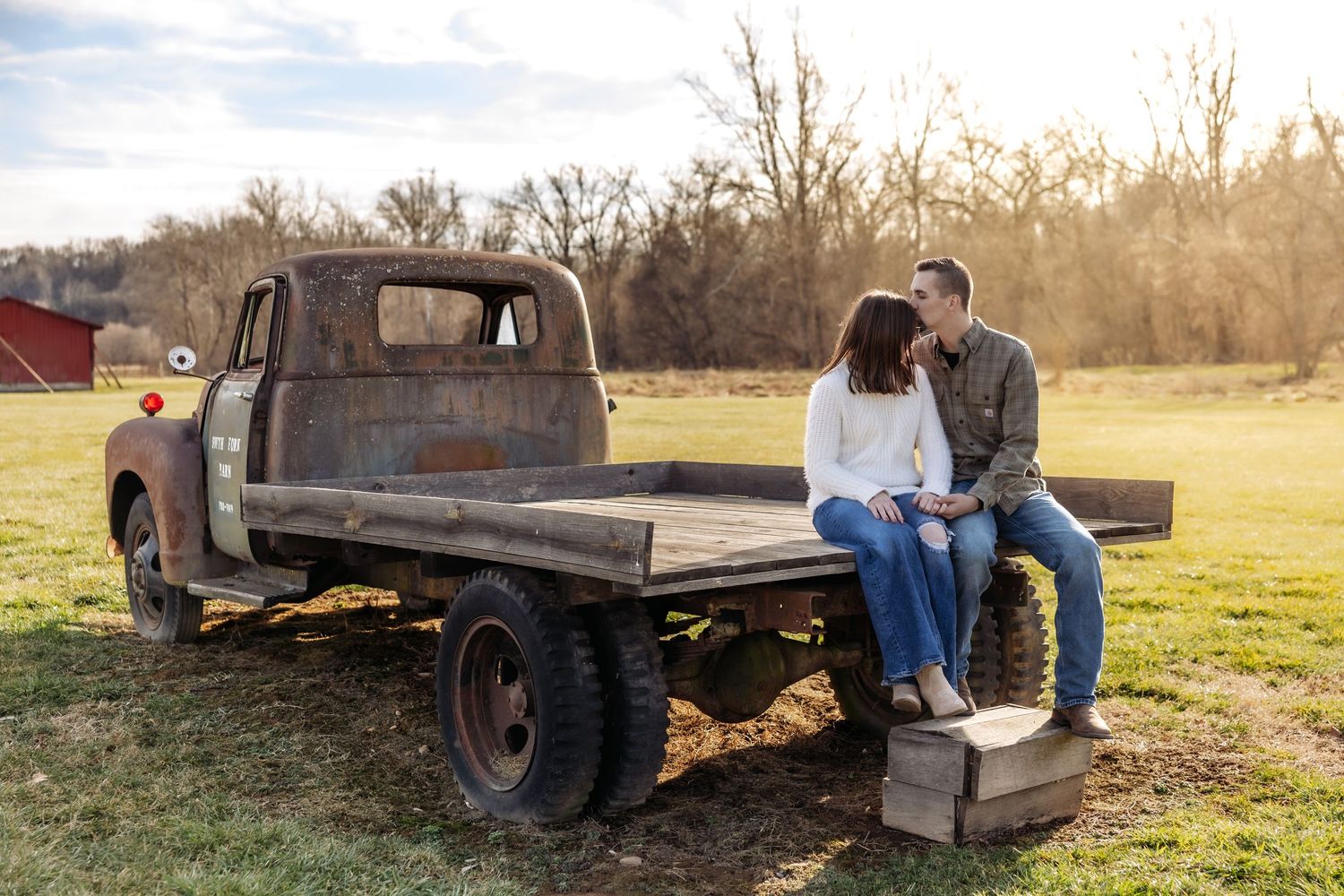 A couple sits together on a vintage flatbed truck in a rustic farm setting during golden hour.