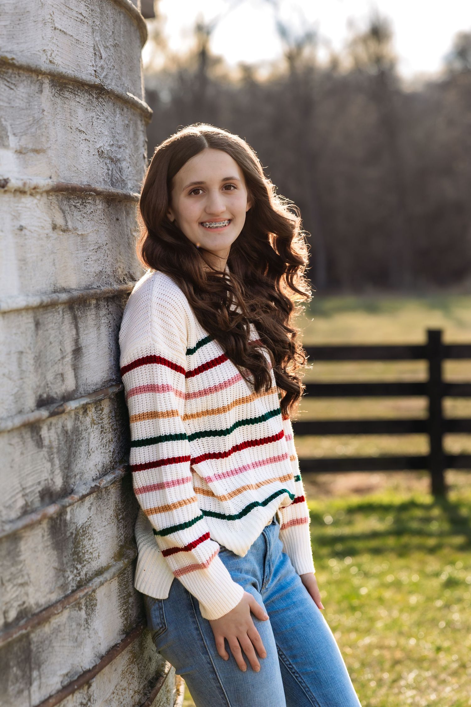 A teenager in a striped sweater and jeans leans against a silo in a rustic outdoor photo with golden sunlight.