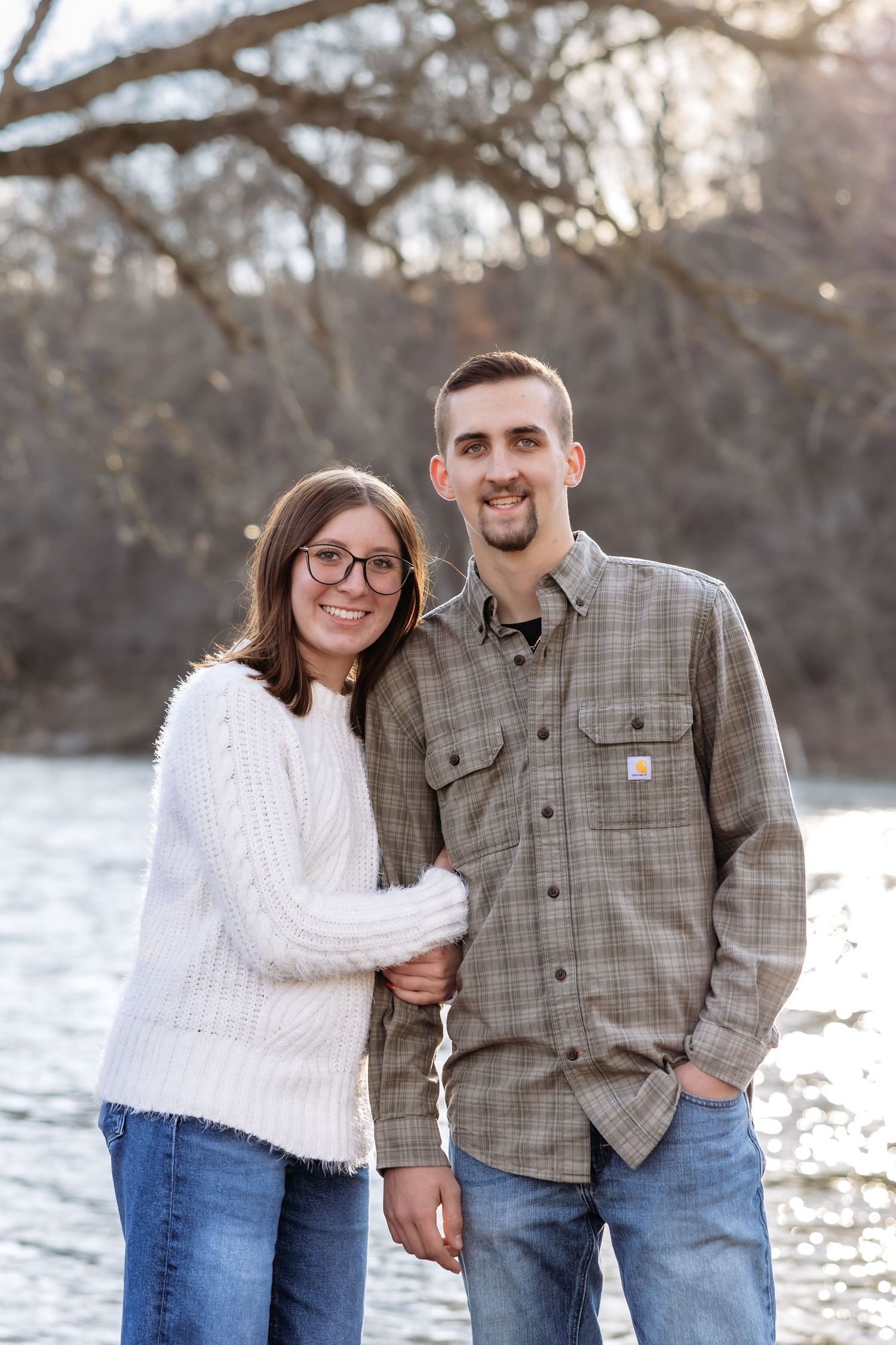 A couple stands together smiling by the Shenandoah River on a winter day with bare trees in the background in the Shenandoah Valley