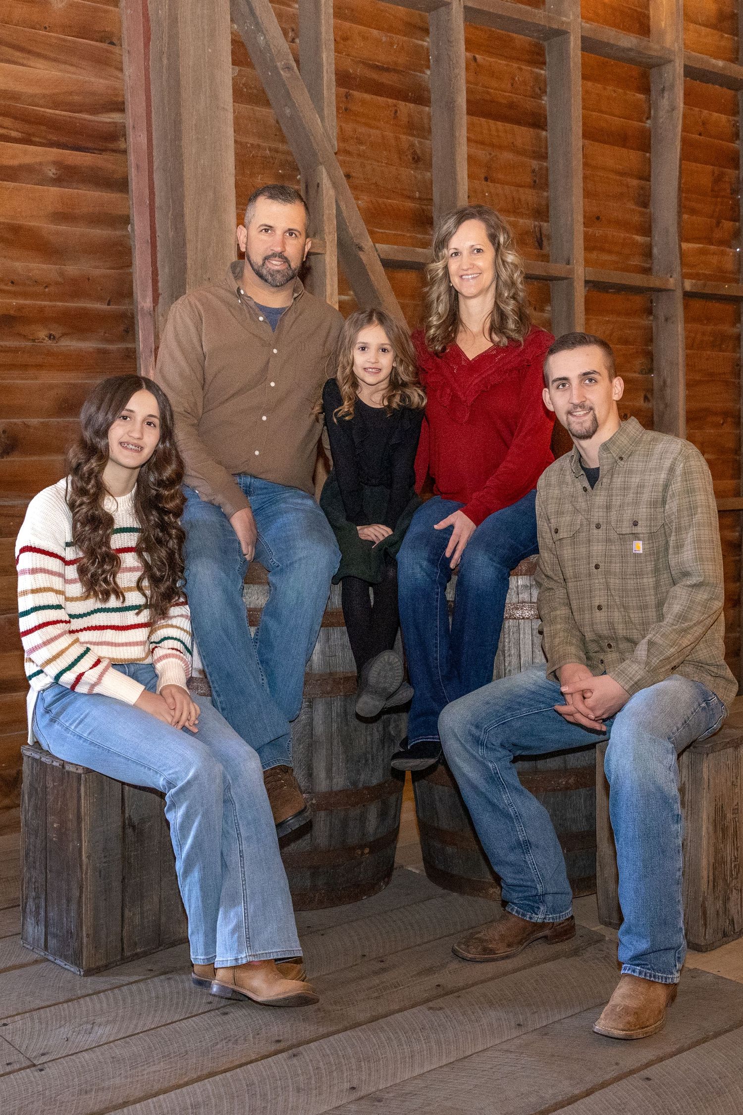 A family poses together on wooden benches inside a rustic barn structure for a casual portrait session.
