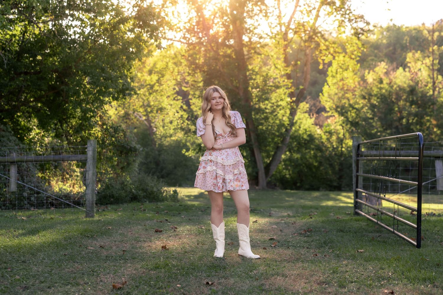 Young woman in floral dress and white boots standing in sunlit green yard near rustic metal gate.