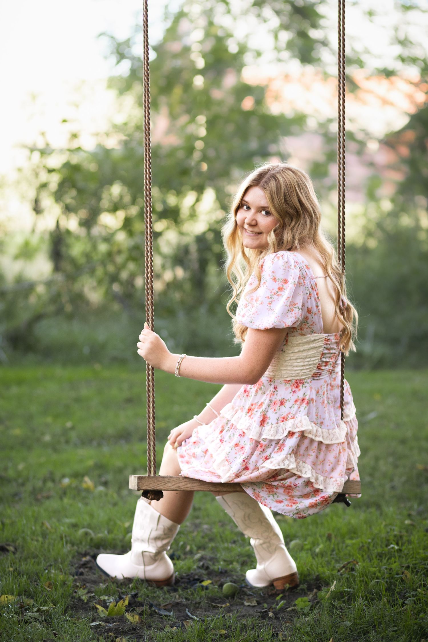 Young blonde woman in floral dress and white cowboy boots sitting on wooden rope swing in sunny garden.