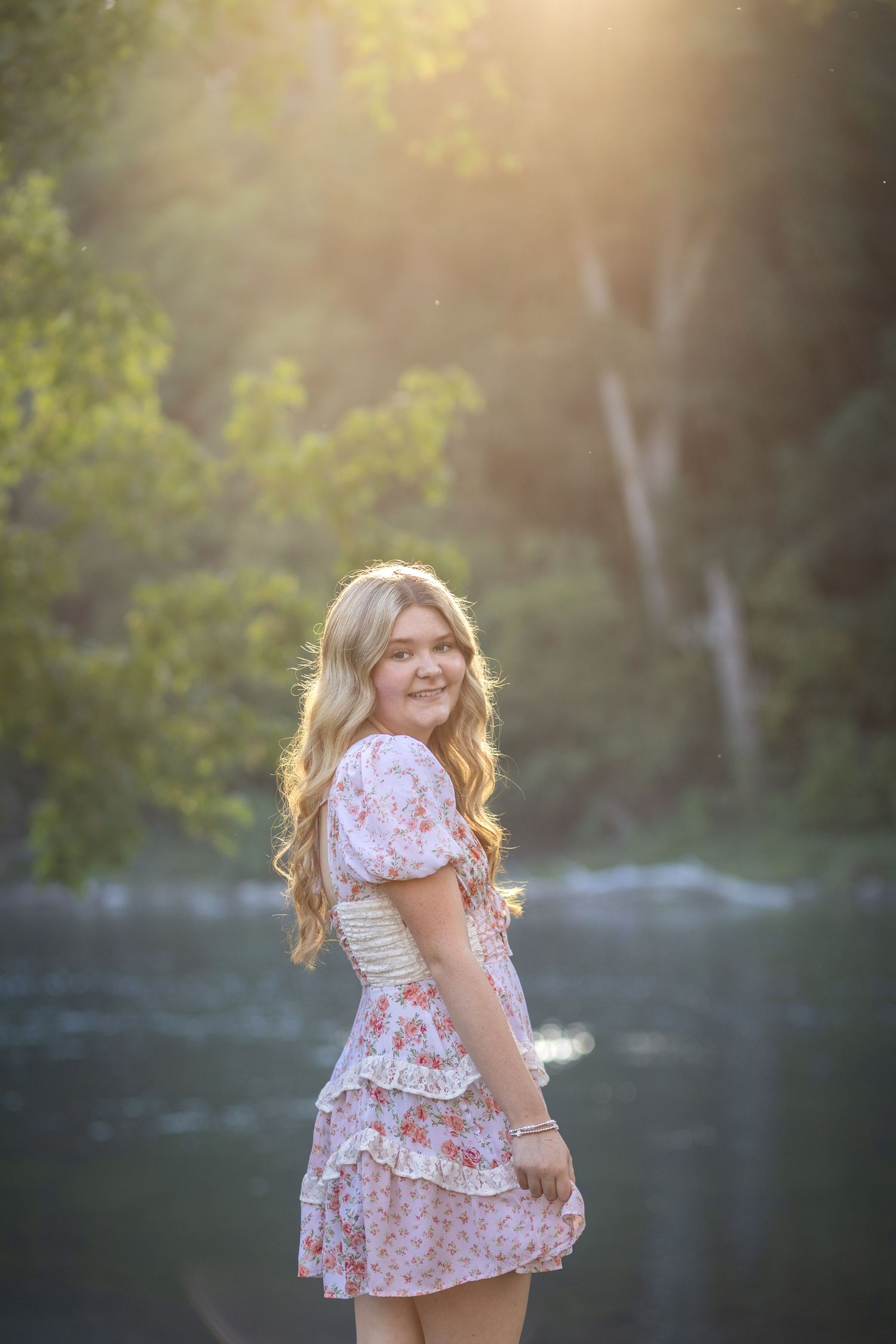 Young blonde girl in floral dress smiling outdoors by river with golden sunlight filtering through trees.