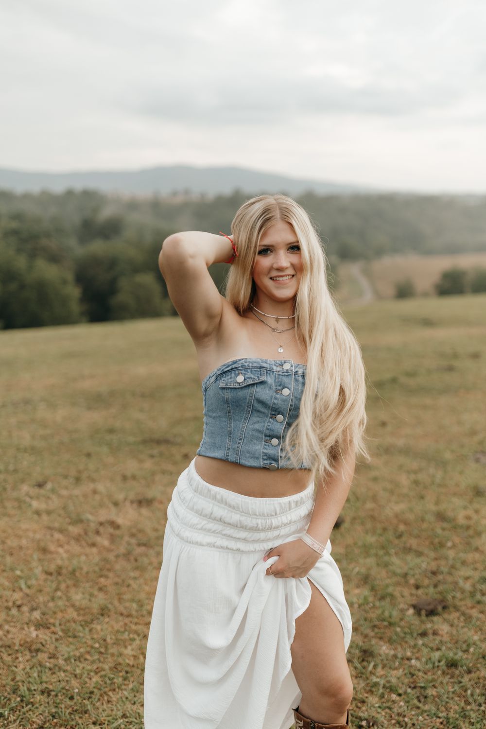 Summer fashion photo of person in denim top and white skirt posing in a grassy meadow with mountains. Virginia graduation/senior session.