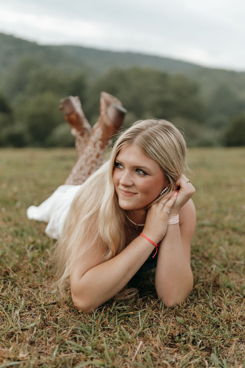 Person with long blonde hair lying in grass wearing boots and white dress in countryside setting. Virginia graduation/senior session.