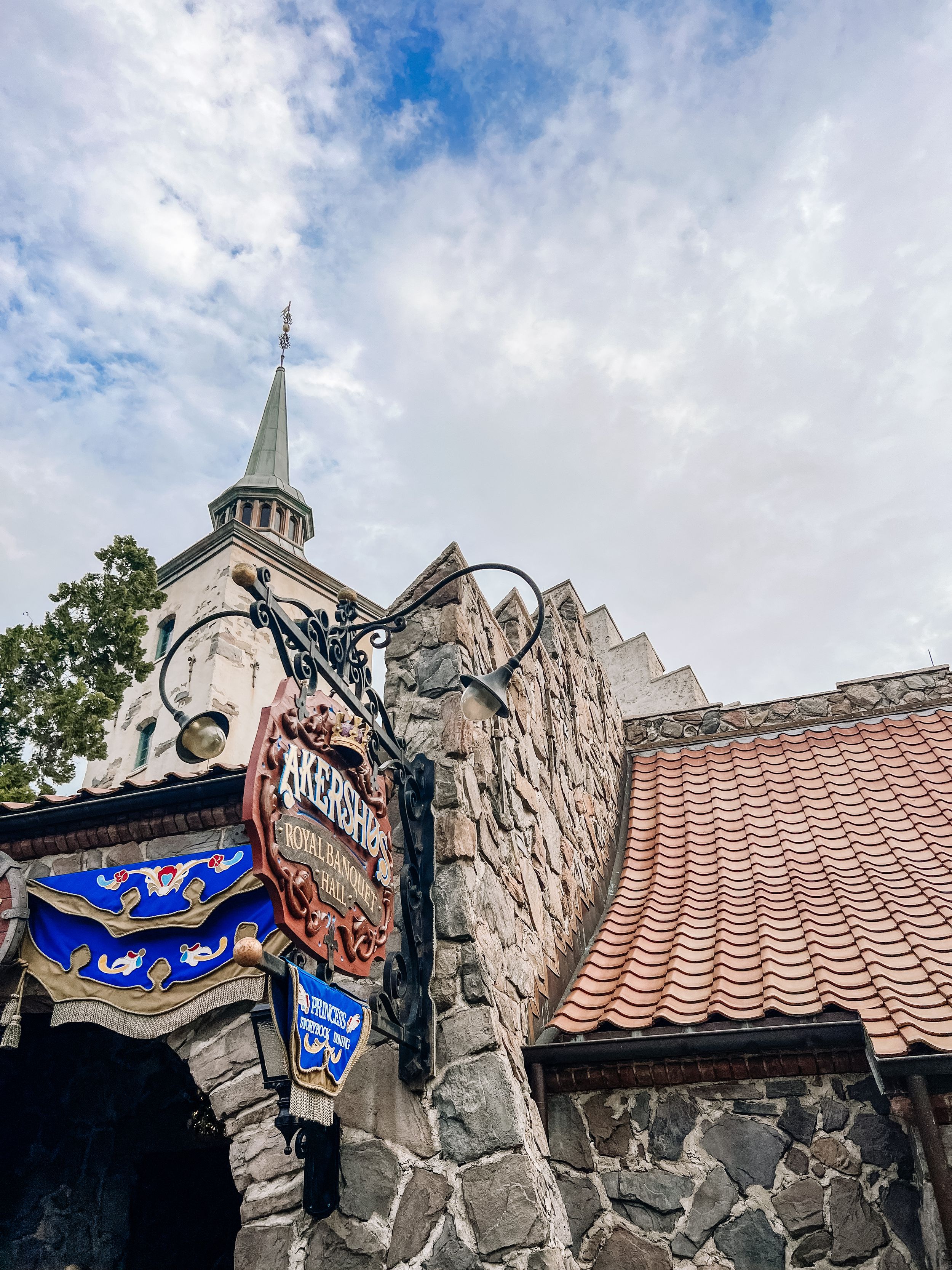 Stone castle facade with ornate signage and spired tower against cloudy blue sky.
