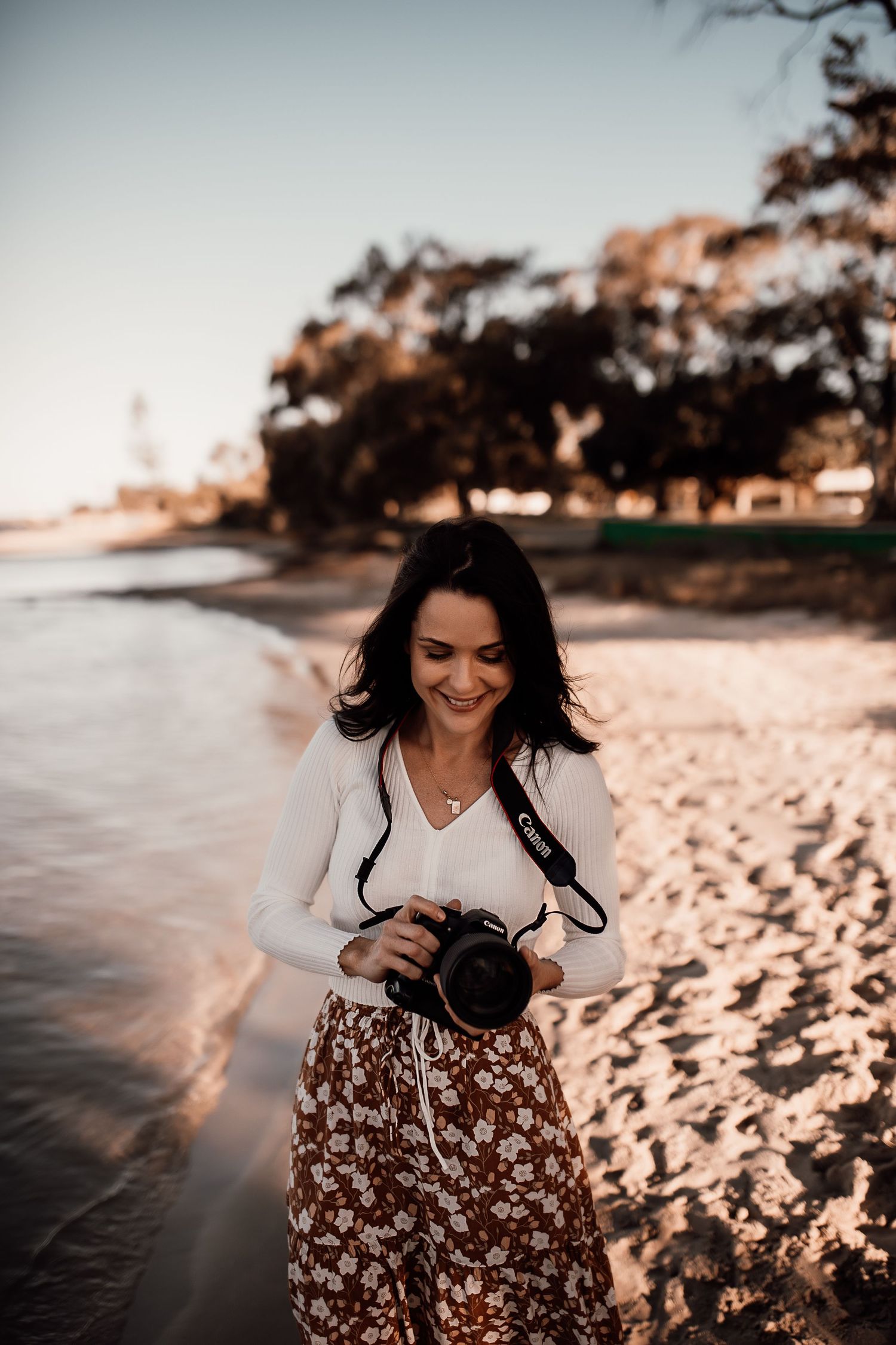 Photographer with long dark hair in white top and floral skirt laughs while holding camera on beach at sunset.