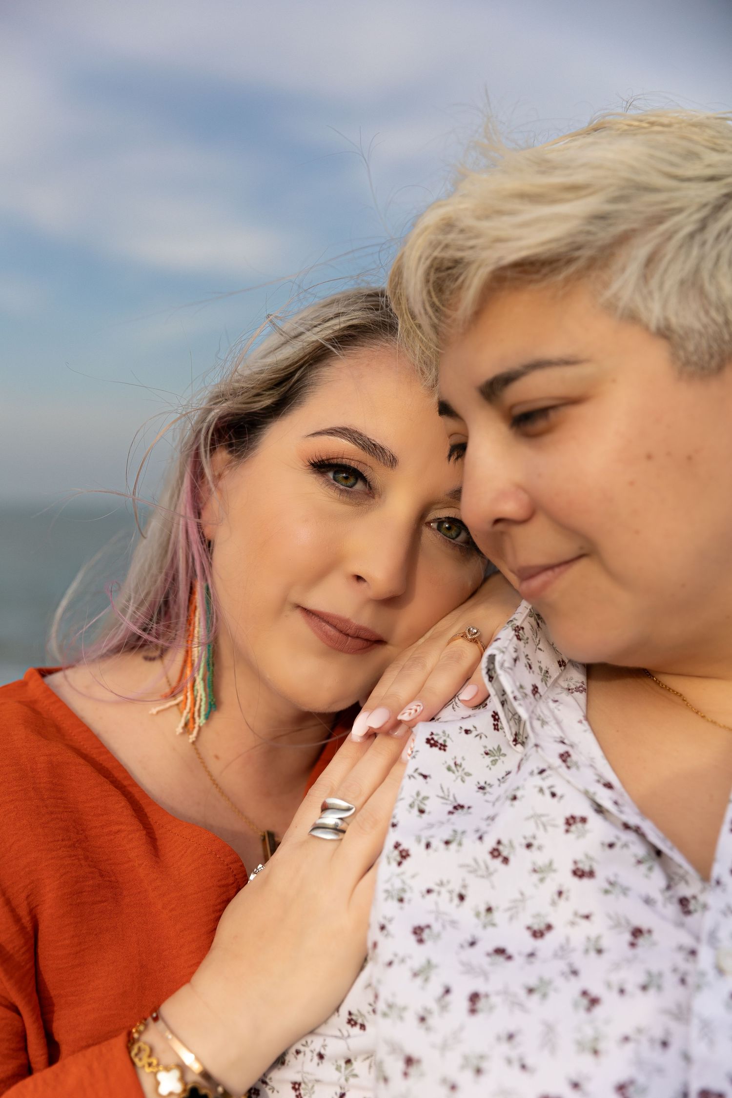 Two people in a romantic close embrace outdoors against a blue sky, wearing an orange sweater and floral top.