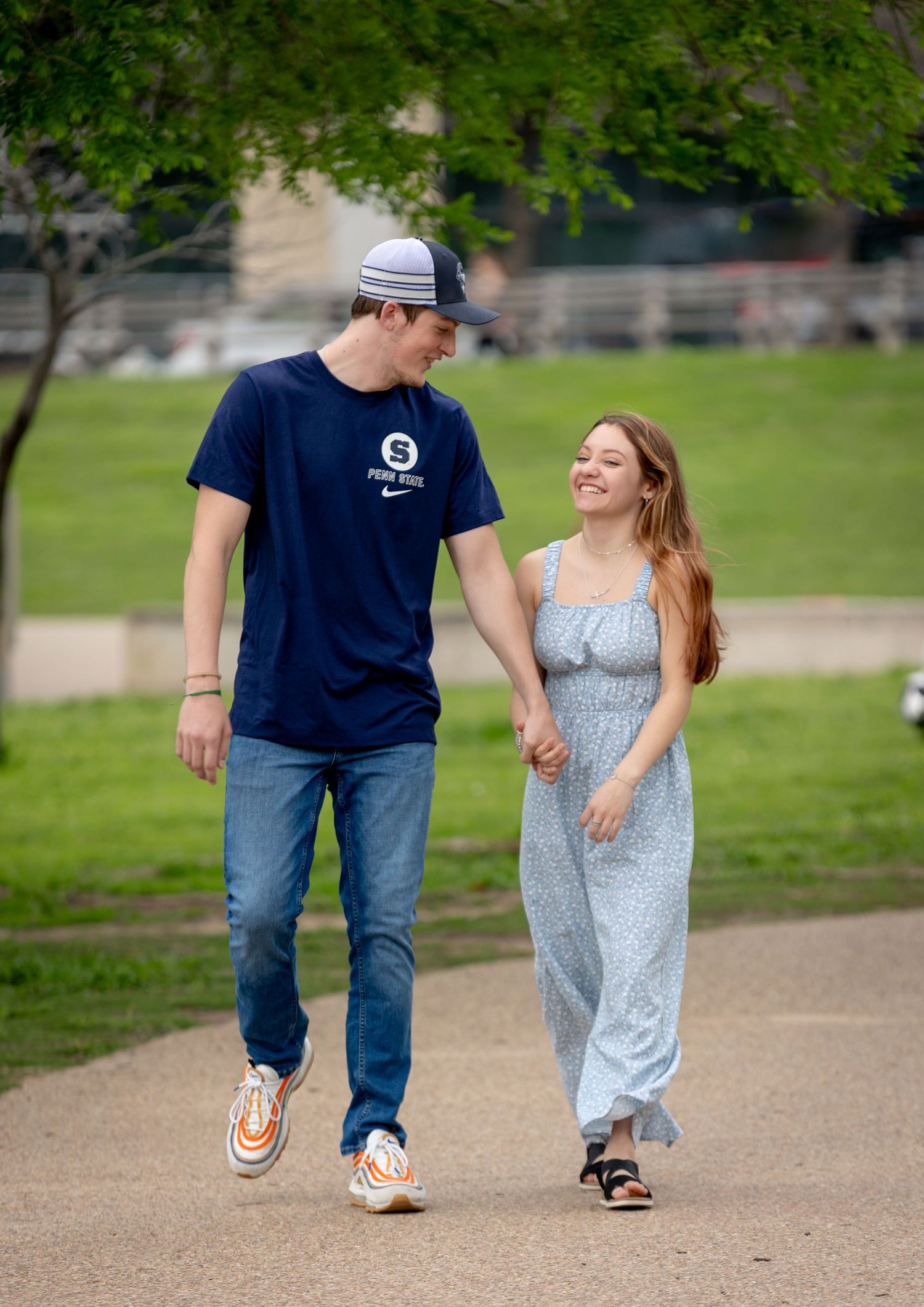 A couple walking together on a path through a park on a sunny day, holding hands and smiling at each other.