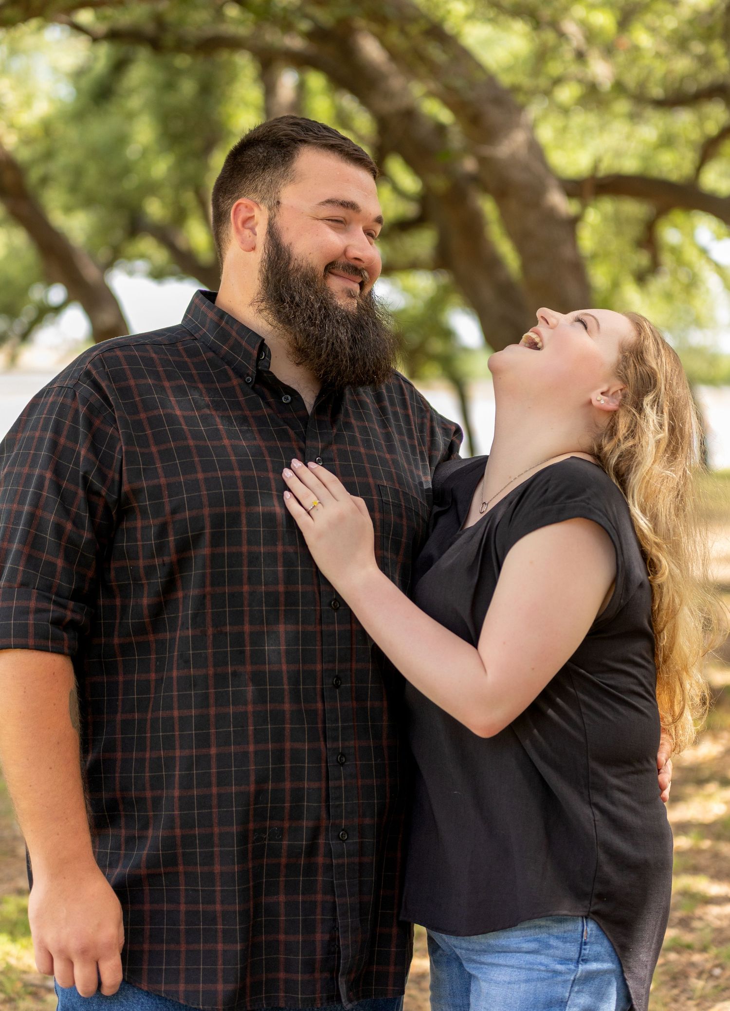 A couple sharing a joyful moment while laughing together outdoors under tree branches.