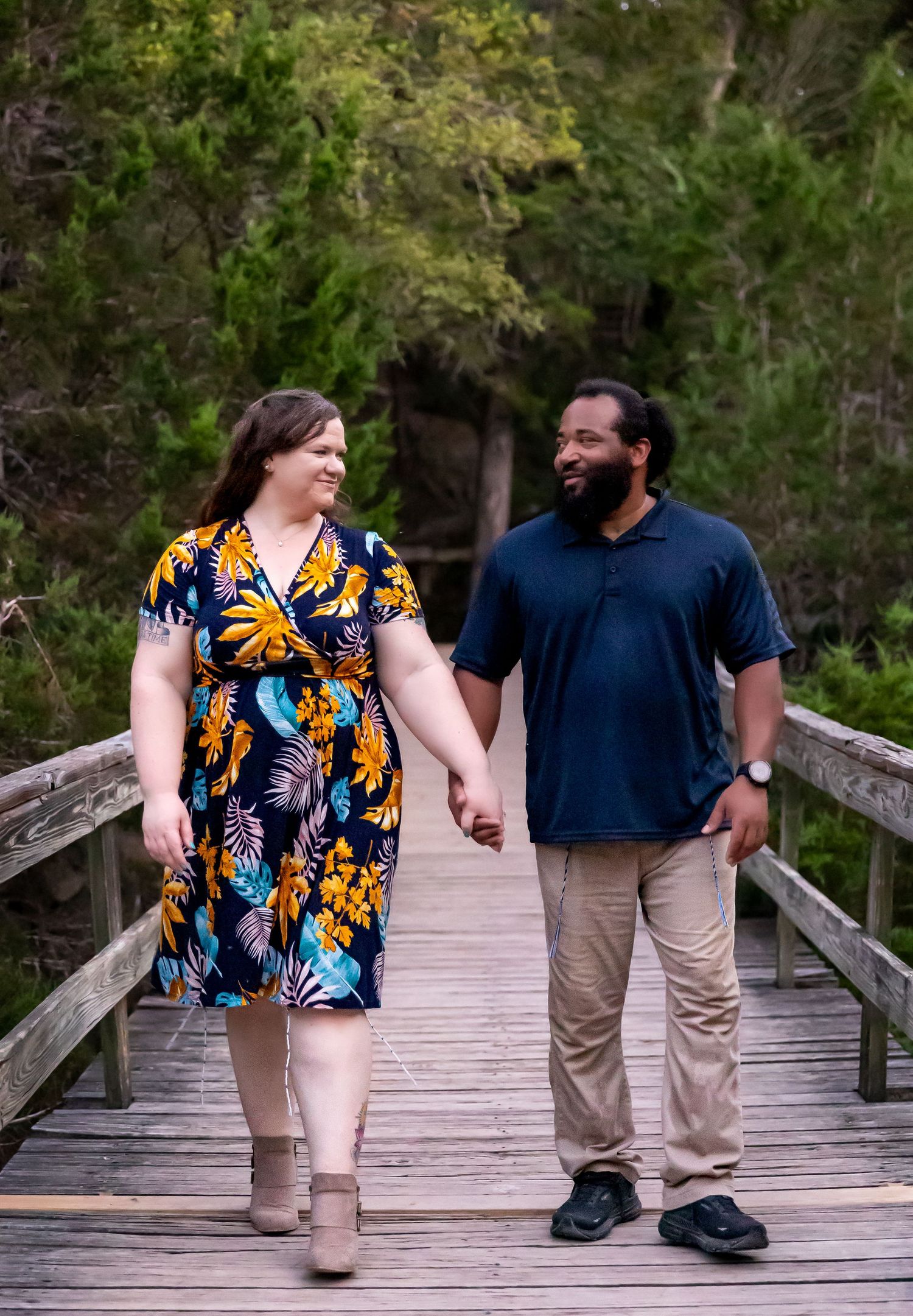 A couple holding hands while walking together on a wooden boardwalk through lush greenery on a sunny day.