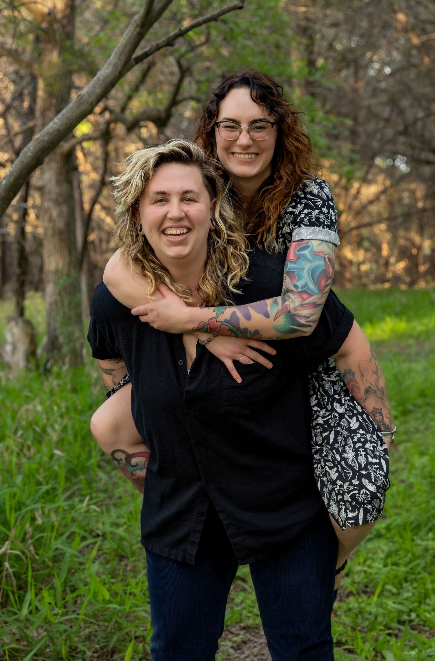 Two people having fun with a piggyback ride outdoors in a wooded area with green grass and trees.
