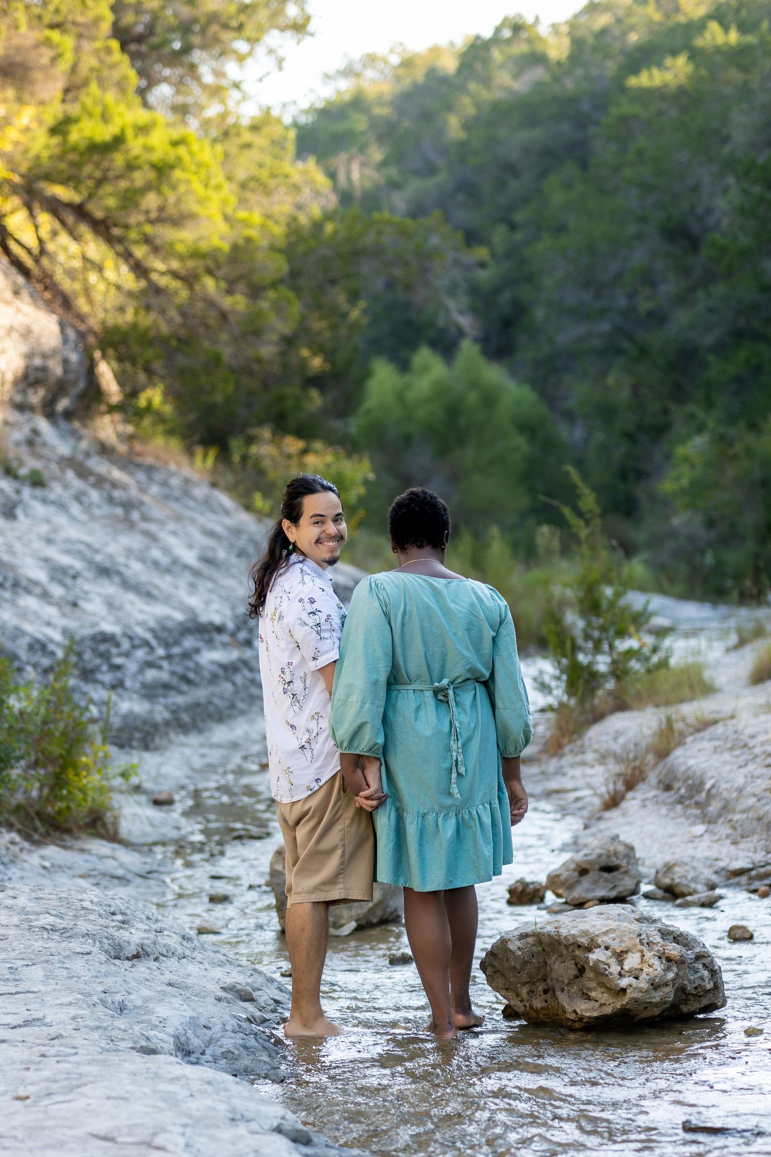 Two people in casual attire explore a rocky creek bed surrounded by lush greenery on a sunny day.