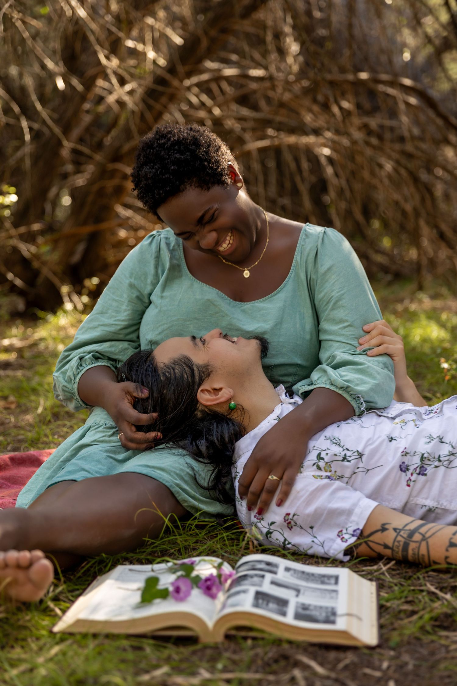 Two people lying in grass reading a book together on a sunny afternoon, wearing turquoise and white clothing.